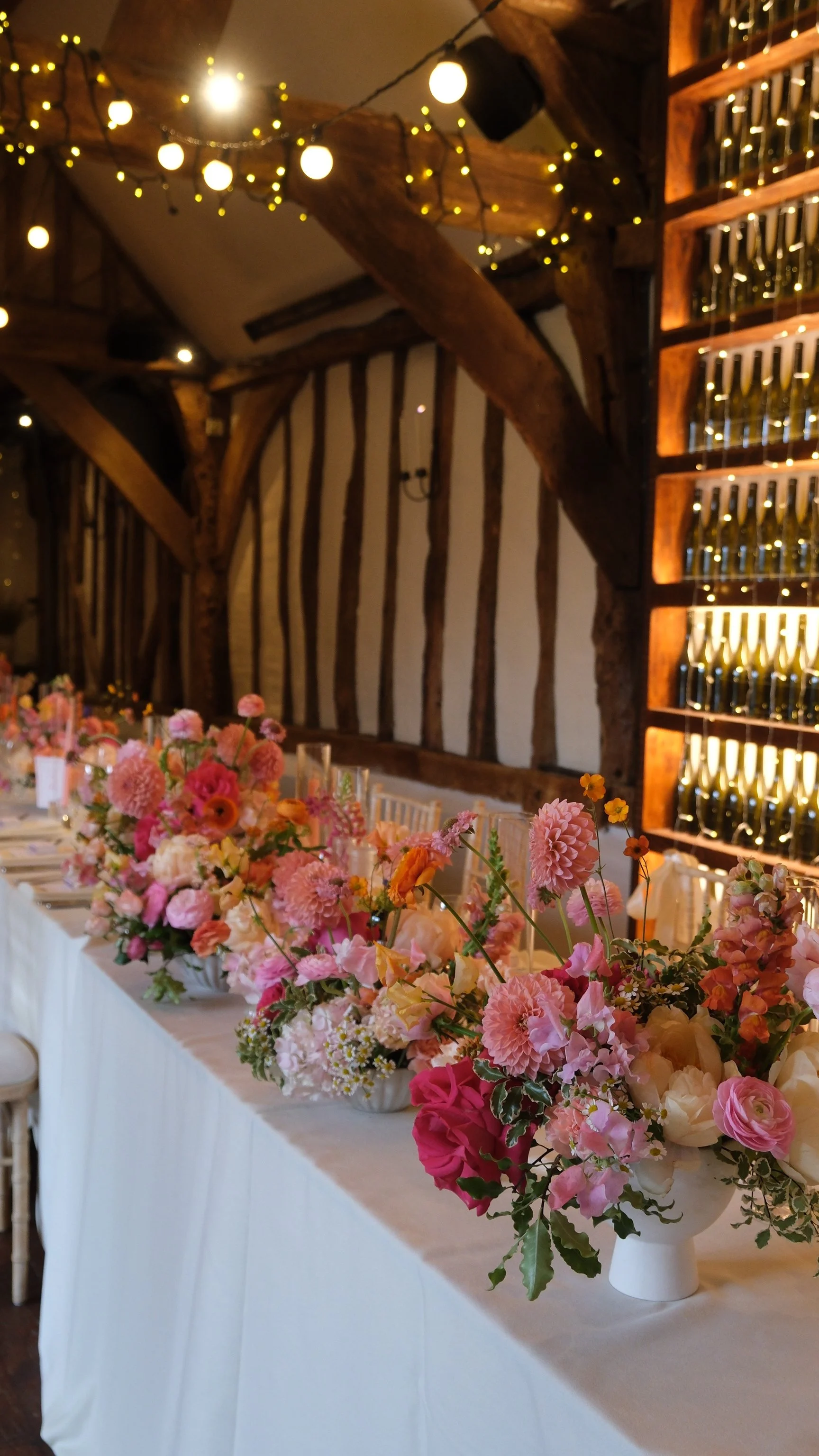 A long table decorated with various pink, white, peach, and orange flowers in small vases, set for a special event, with fairy lights strung on the ceiling above and bottles displayed on a shelf on the wall to the right.