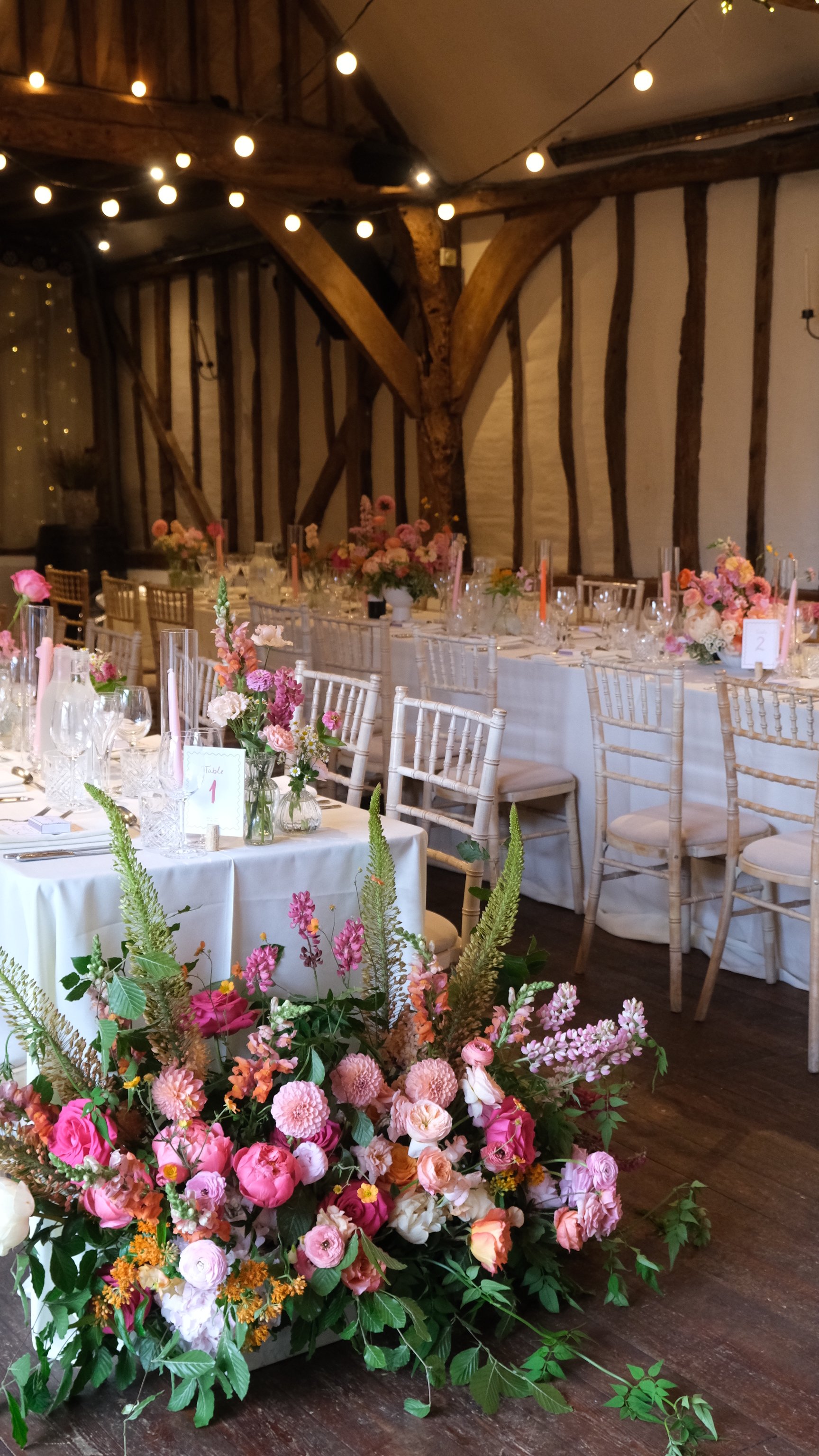 A decorated indoor event space with long tables covered in white tablecloths and tall floral centerpieces. The room features exposed wooden beams and string lights, creating a warm and rustic atmosphere.