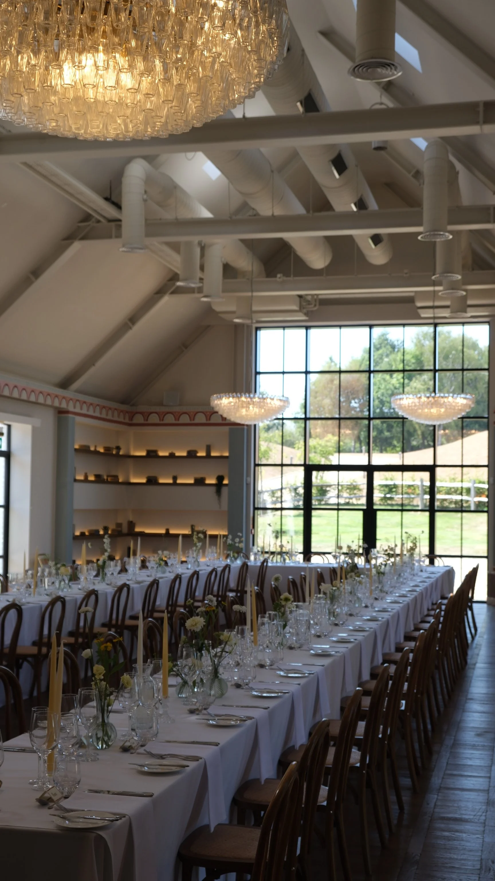Elegant banquet hall with long dining tables decorated with white tablecloths, floral centerpieces, and glassware. Large windows let in natural light, and modern chandeliers hang from the ceiling.