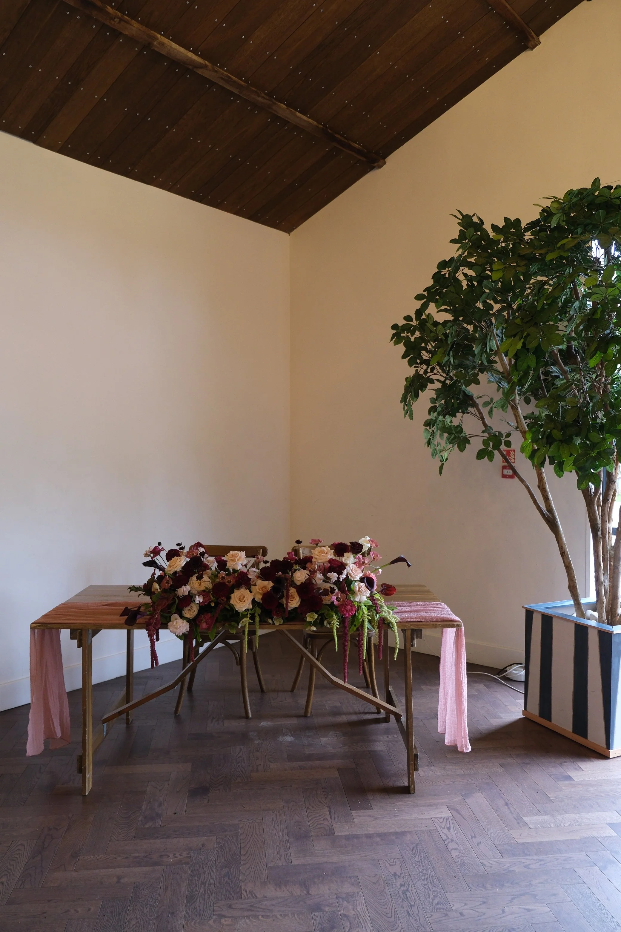 A wooden table covered with a pink tablecloth and a large floral arrangement consisting of pink, cream, and deep purple flowers with green foliage; a wooden ceiling and wooden floor, with a large potted plant to the right and a light-colored wall in the background.