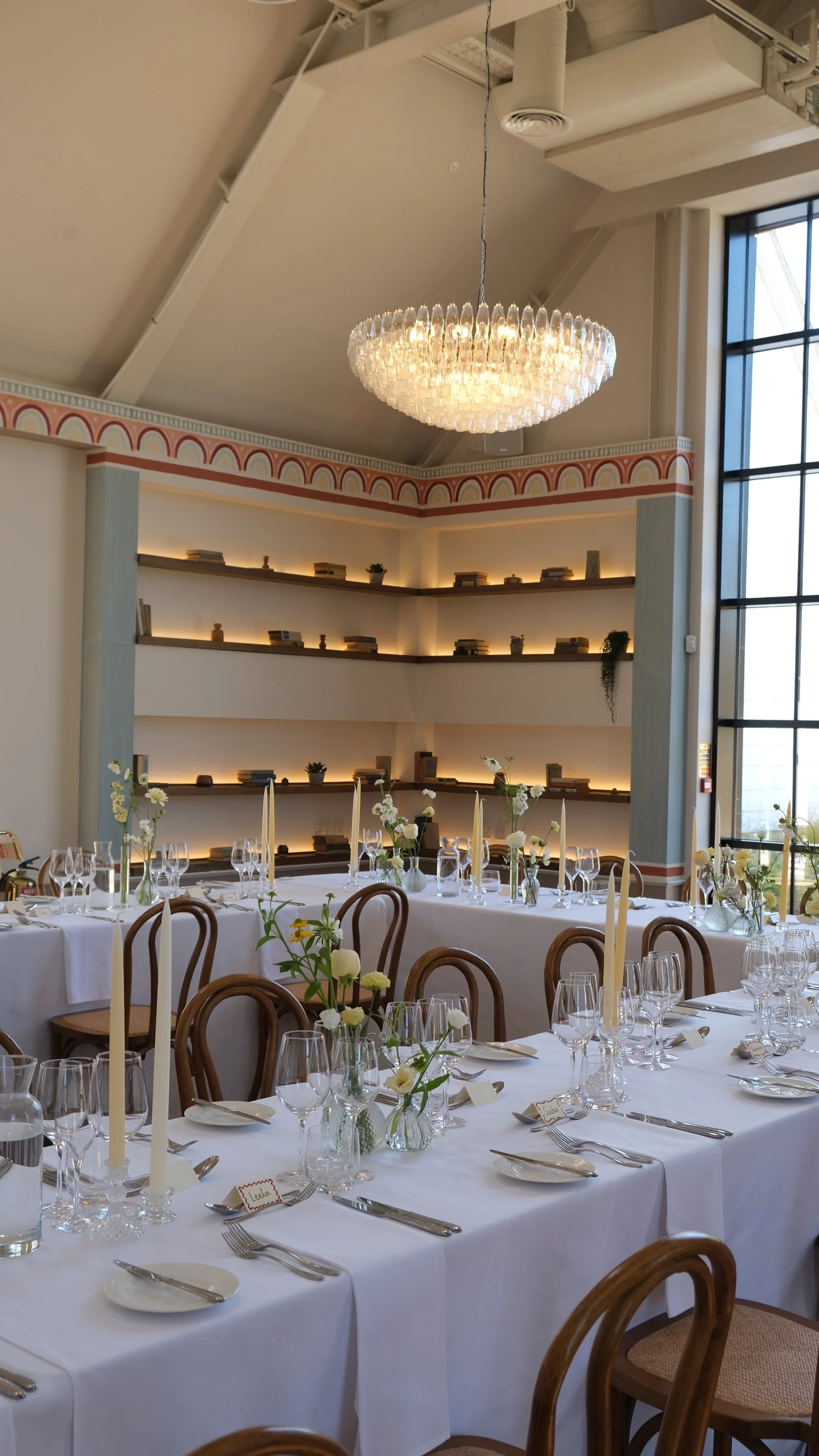 Elegant dining room set for a wedding or special event, featuring long white tables with glassware, candles, and floral centerpieces, surrounded by wooden chairs. A chandelier hangs from the ceiling, with built-in lighting on a wall with bookshelves and decorative plants. Large windows allow natural light into the room.