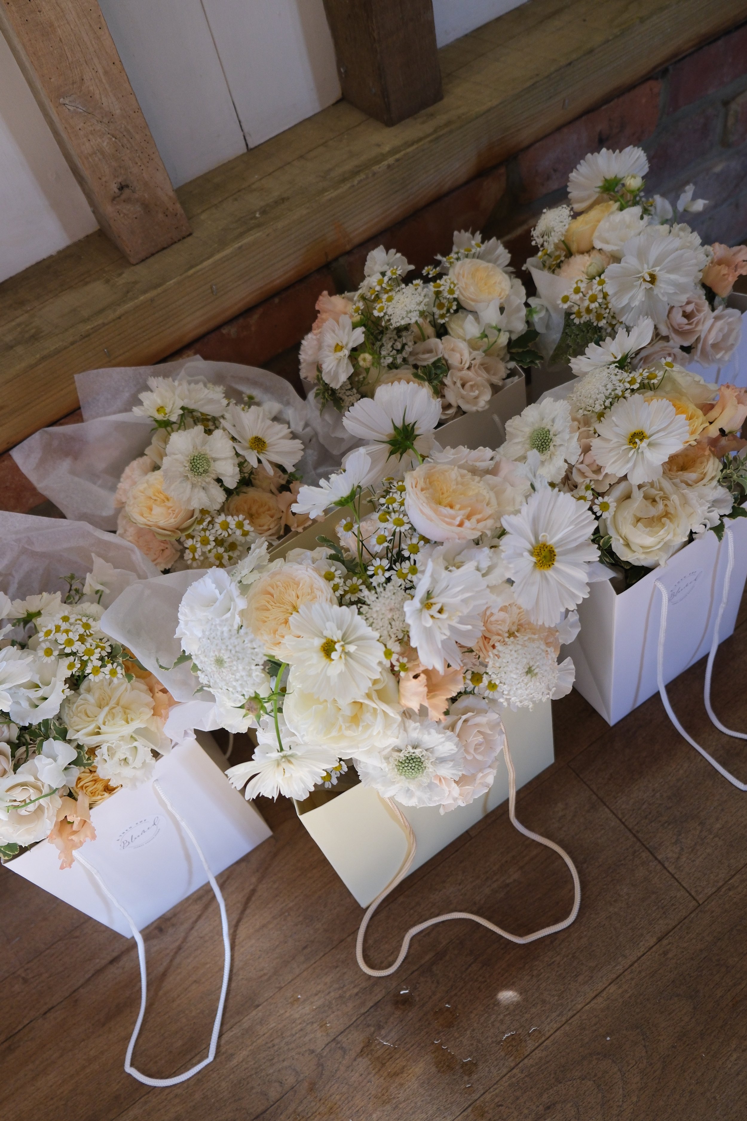 Several bouquets of cream and white flowers are placed in white gift bags on a wooden floor, against a brick wall with a wooden shelf above.