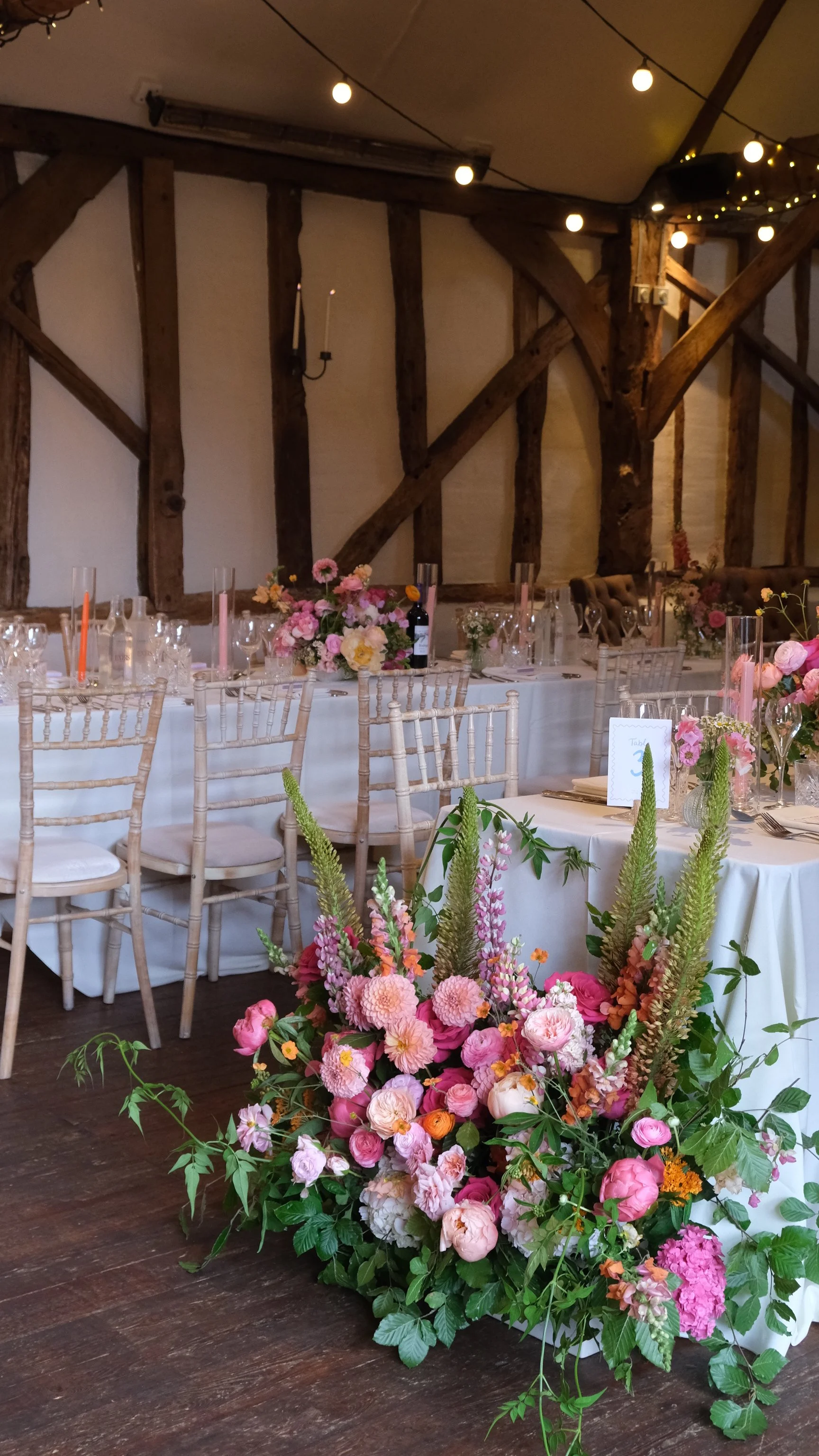 A decorated indoor event space with wooden beams, a long table with floral centerpieces, and a large flower arrangement in the foreground featuring pink, orange, and peach flowers and green foliage.