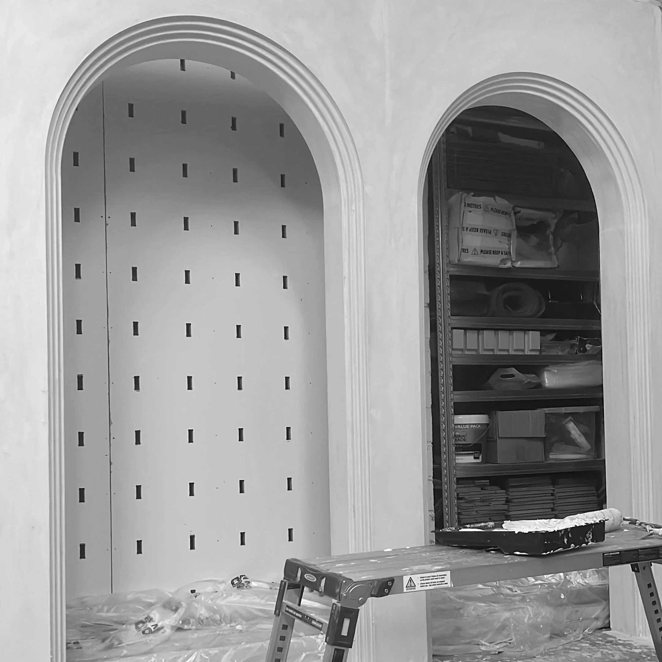 Interior view of an unfinished room with two arched doorways, construction tools on the floor, and storage shelves filled with building supplies and tools.