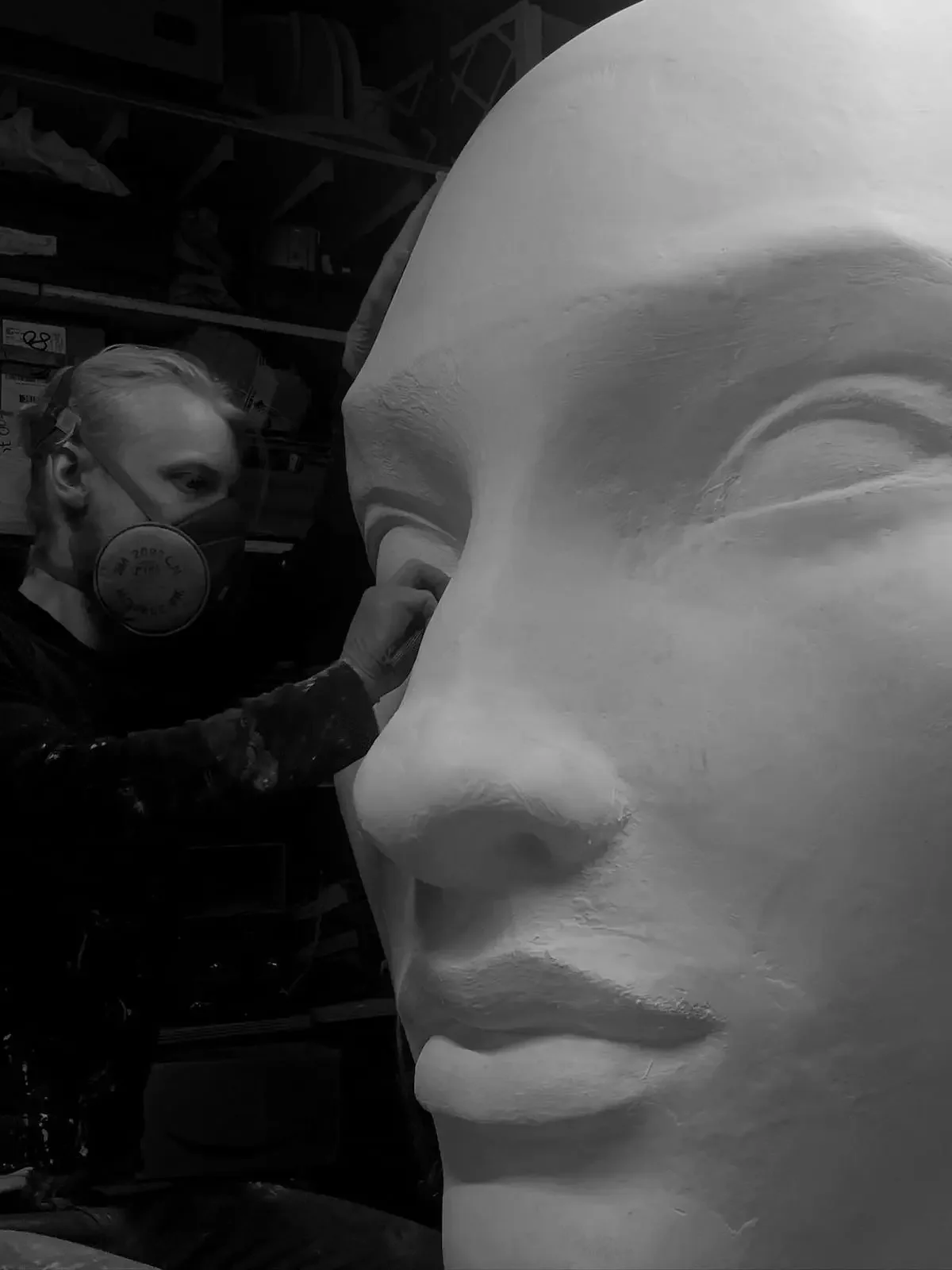 A person wearing a mask and protective eyewear working on a large sculpture of a face, possibly in a studio or workshop.