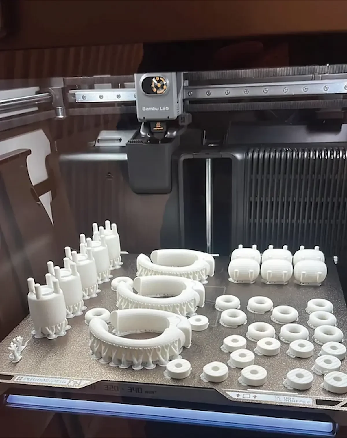 3D-printed white plastic mechanical components arranged on a work surface beneath a 3D printer.