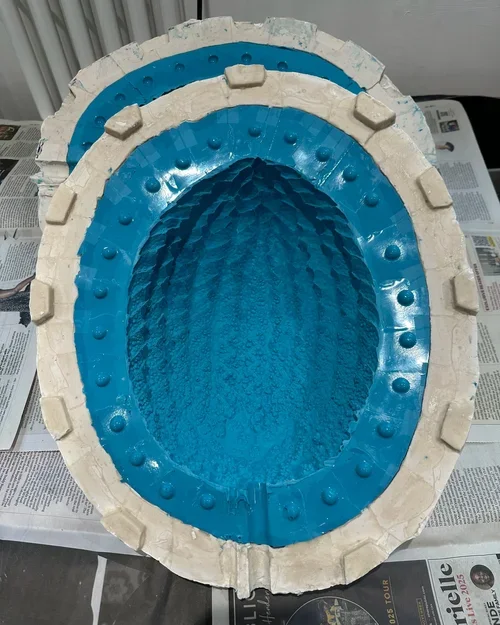 A large, oval-shaped object with a blue interior and a beige exterior, placed on top of newspapers. The inner surface has a textured pattern, and the outer edge features evenly spaced rectangular protrusions.