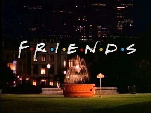 A sign spelling 'FRIENDS' with colored dots between the letters, displayed over a fountain and a building at night.