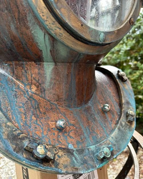 Close-up of a rusty, weathered metal object with bolts and a circular glass window, outdoors with green foliage in the background.