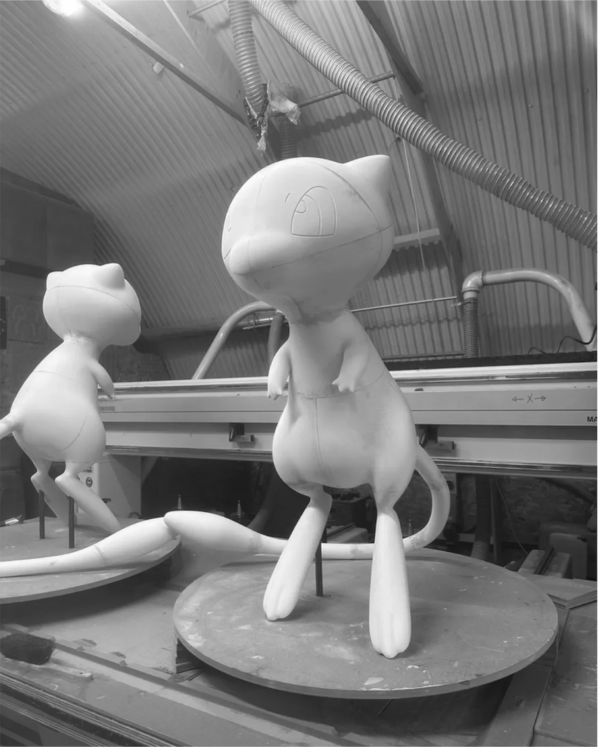 Black and white photo of cartoon-like cat sculptures in a workshop, one standing on its hind legs with arms out and a smiling face, and the other leaning forward with one arm raised.