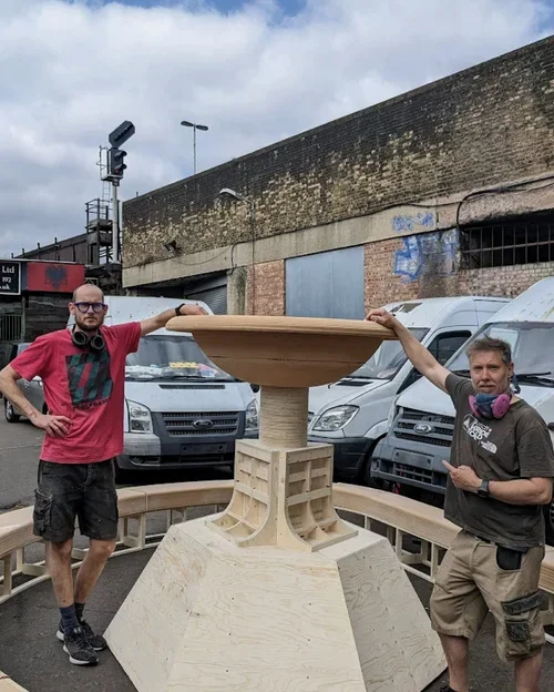 Two men with headsets stand beside a large, unfinished wooden fountain structure outdoors, with vehicles and a brick wall in the background.