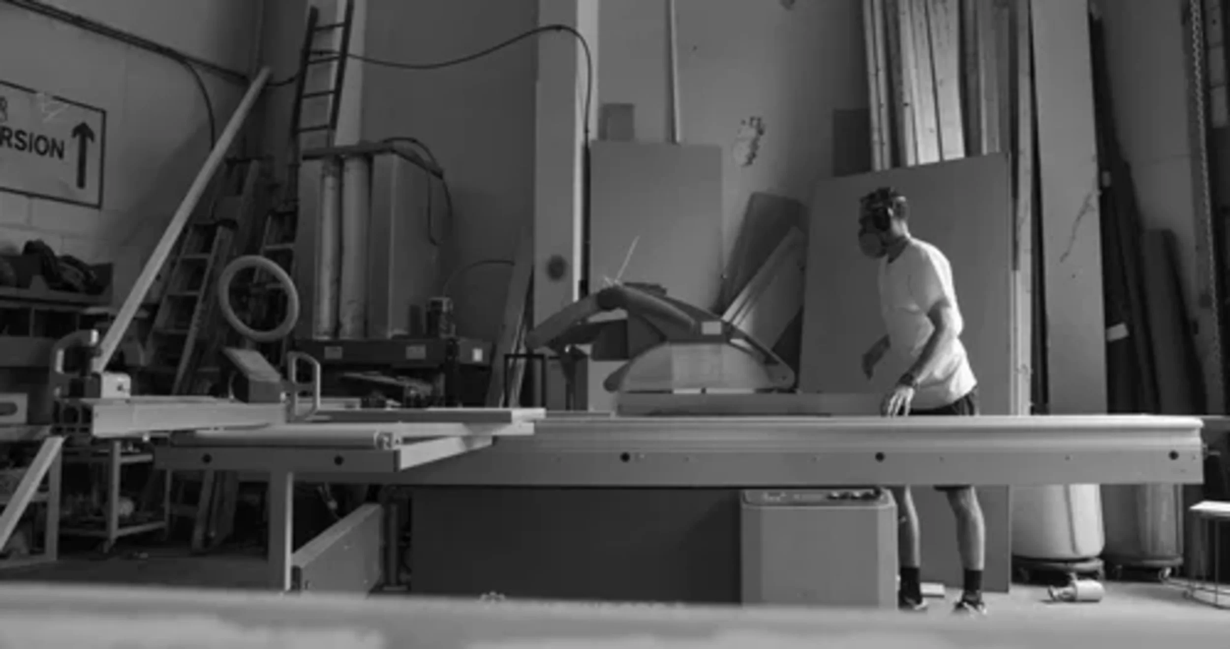 A person wearing safety goggles and a mask working in a woodworking shop with various tools and equipment, including a large saw and wooden materials.
