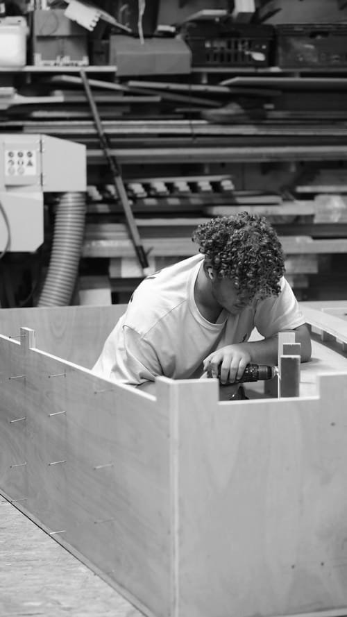 A young man with curly hair and light-colored t-shirt working with a power drill on a wooden piece in a woodworking shop. Shelves with various wooden planks and tools are in the background.