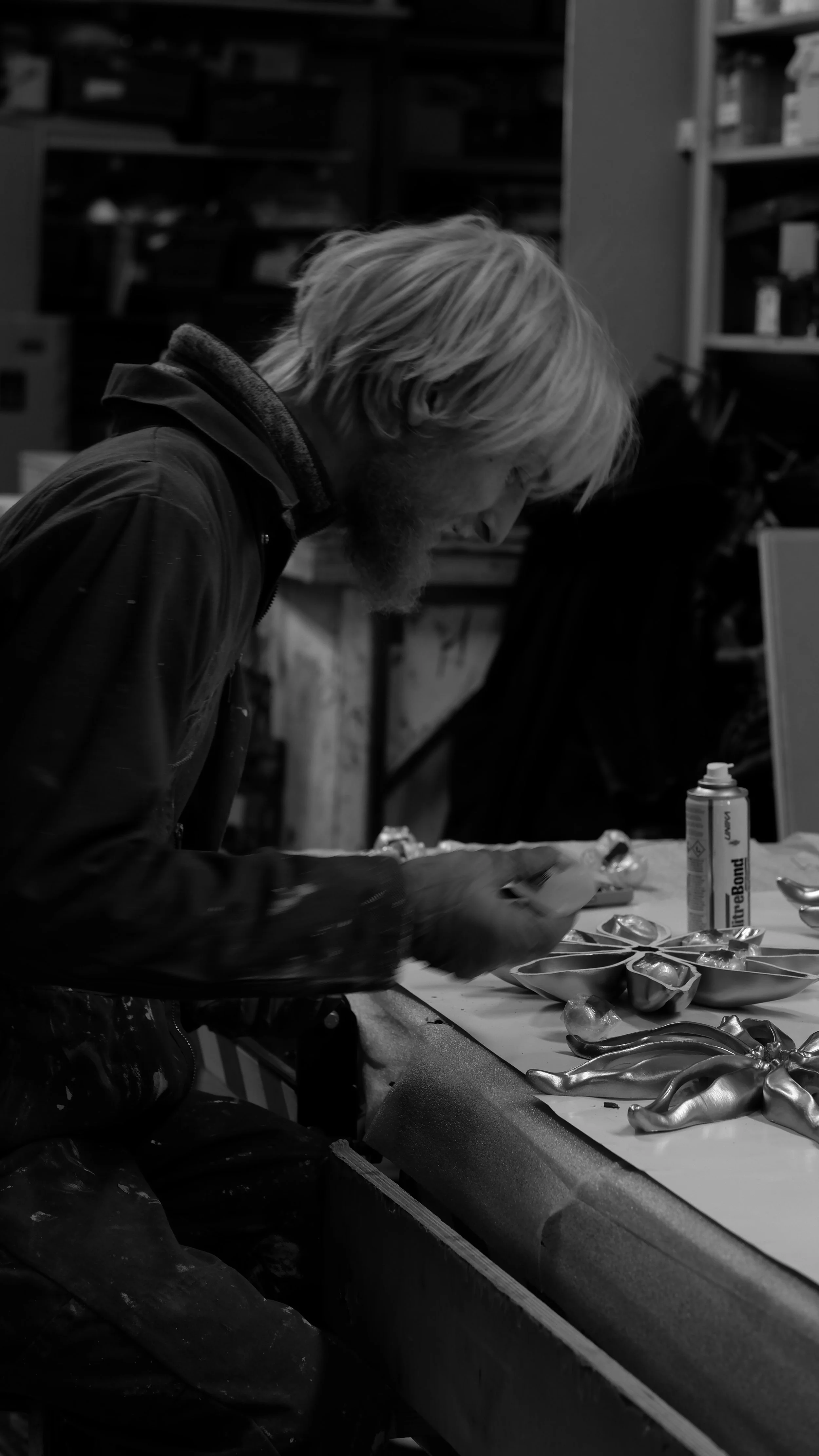 A man with long hair and a beard working on a project in a workshop, with various tools and a can of spray paint on the table.