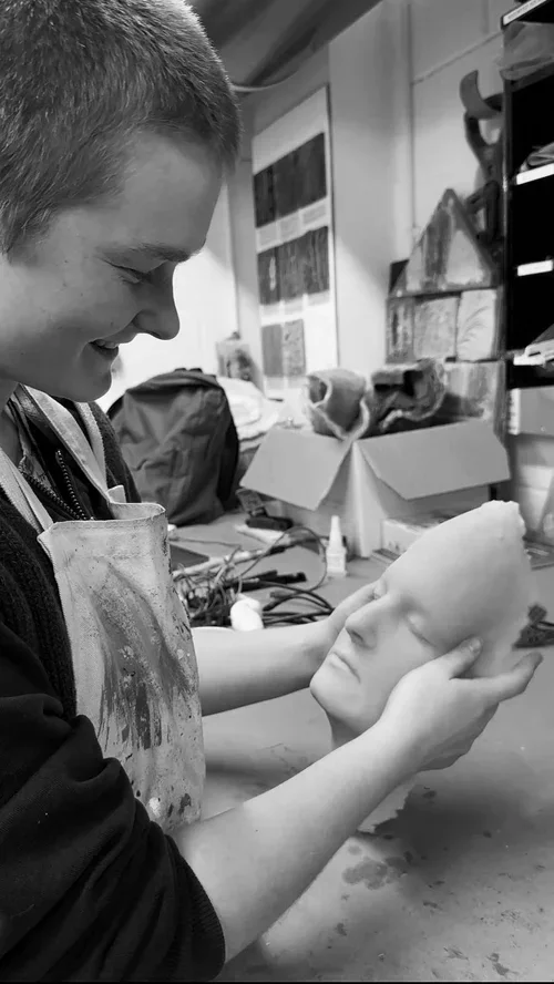 A happy prop maker holding a realistic lifecast of their face in a workshop/ art studio.