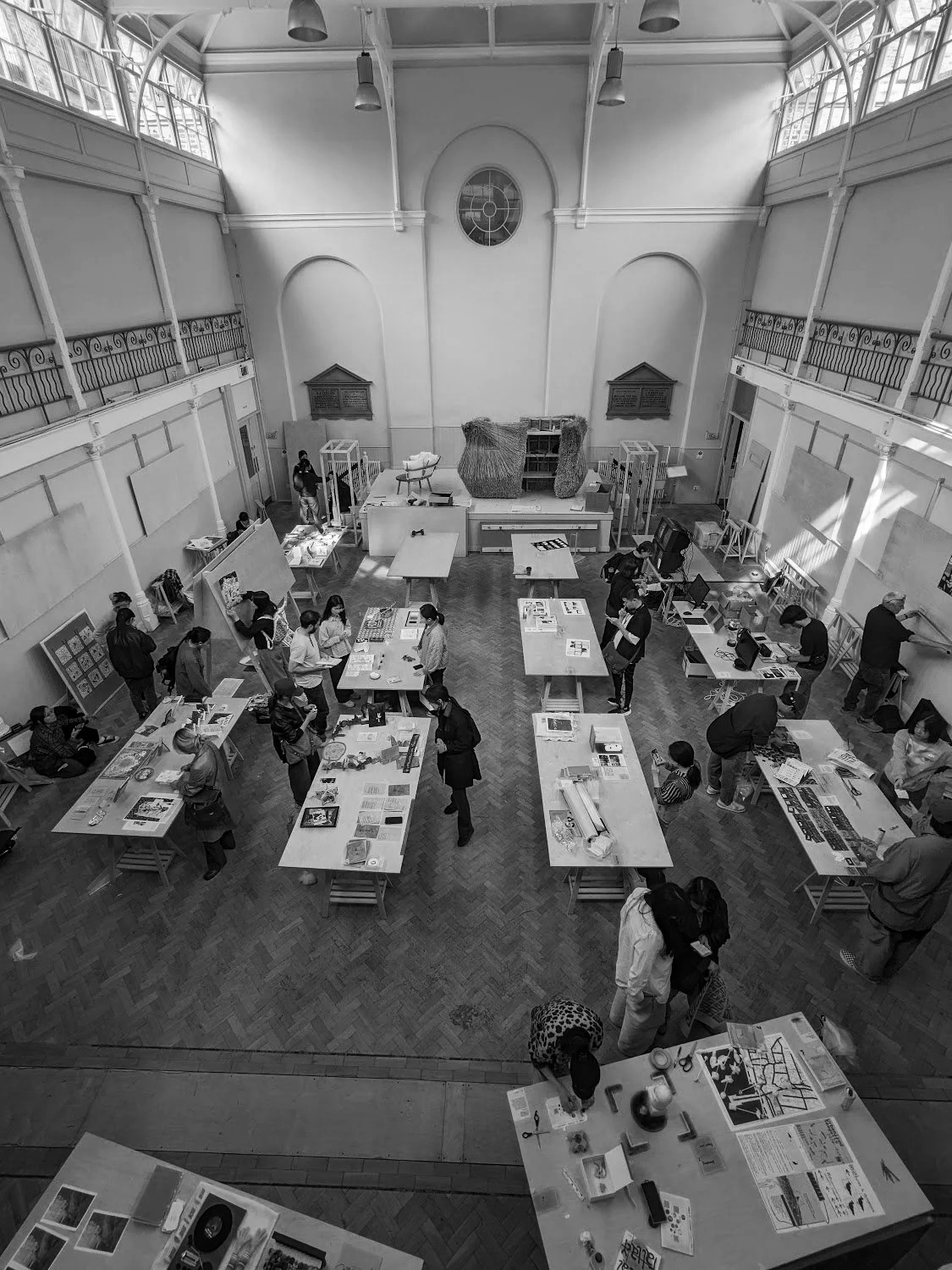 Black and white photo of a spacious event hall with tables displaying artwork and craft projects, and people browsing and talking.