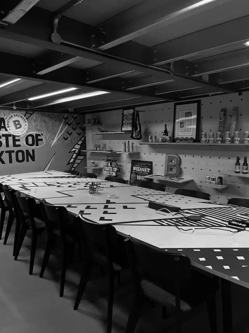 Inside the Brixton Brewery taproom with a long table, shelves with canned drinks, framed picture, and decorative items, all in black and white.