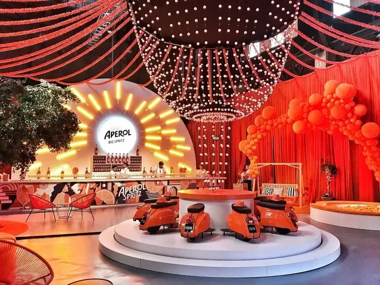 Decorative event space with a stage featuring a large illuminated sign that reads "Aperol Big Spritz" with a bar, orange and red balloons, sofas, and scooters on a circular platform, with red curtains and a canopy of string lights overhead.