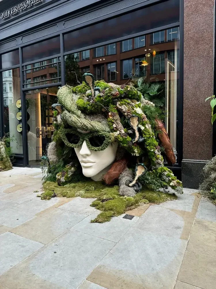 A large outdoor art installation of a woman's face wearing sunglasses, with her hair made of moss, plants, and natural elements, placed outside a building with glass windows.