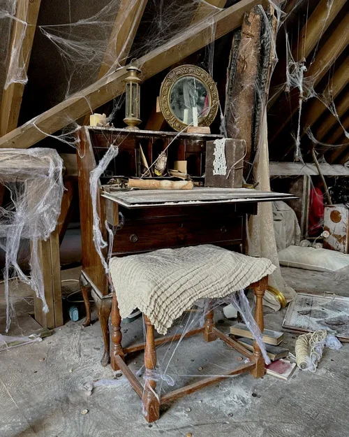 An old wooden desk with a shelf on top, covered in dust and spider webs, in an attic or basement. The top shelf has a mirror, candles, and various small items. The desk has a blanket draped over the front and scattered debris on the floor.