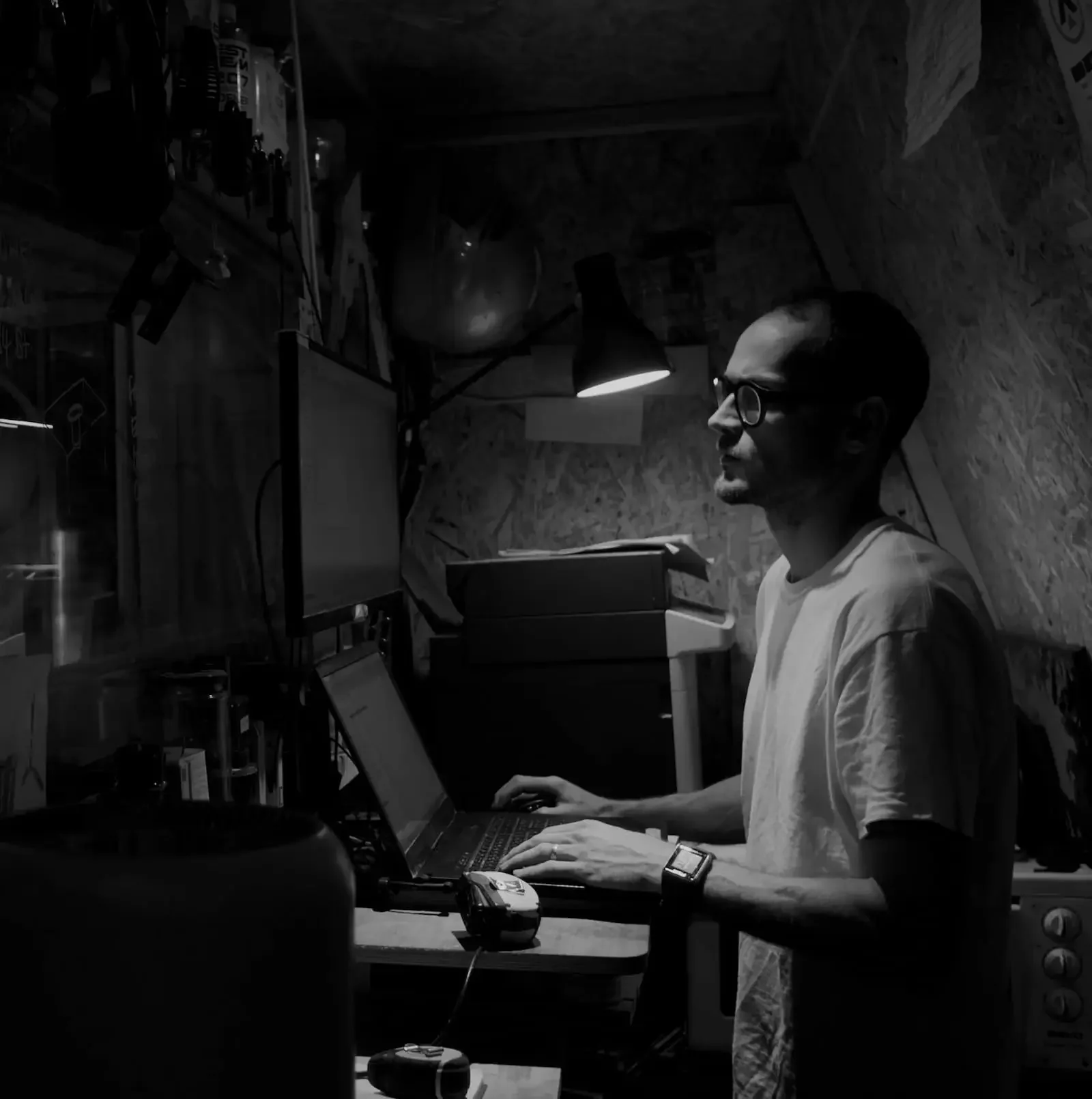 A man wearing glasses and a watch working on a laptop in a dimly lit room with various electronic equipment and shelves in the background.