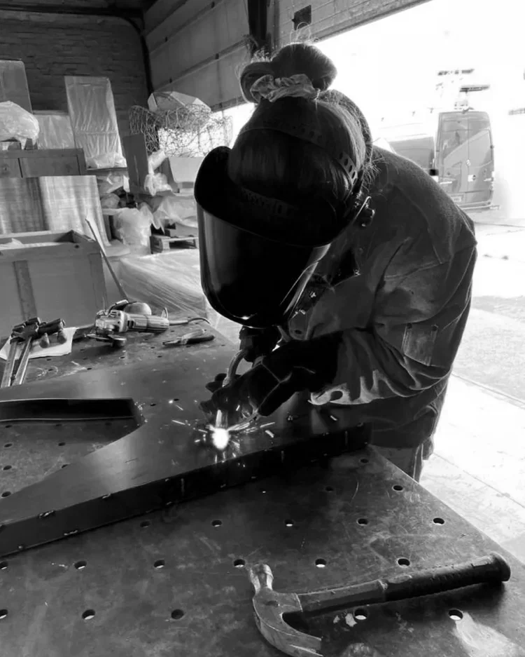 Person welding metal in a workshop, wearing a protective helmet and gloves, with various tools and materials on the workbench.