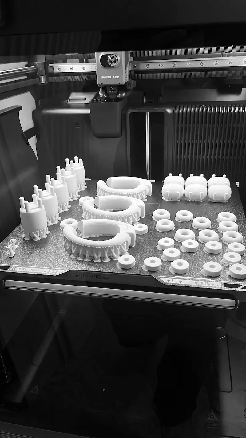 3D printer printing white plastic parts, including rings and curved shapes, on a work surface with various components arranged for fabrication.
