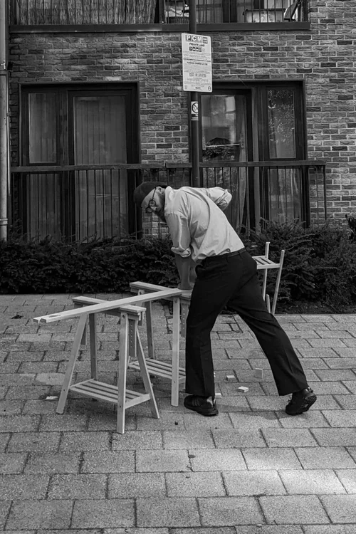 A man wearing a cap, glasses, and light-colored shirt, working on a wooden project outdoors in front of a brick building with windows, using a saw on a makeshift workbench.