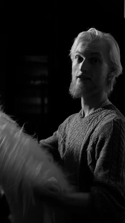 Black and white photo of a man with light-colored hair and beard, wearing a textured sweater, sitting indoors with a dark background.