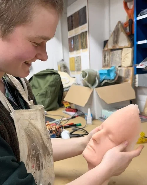 A person holding a realistic sculpture of a baby's head, looking at it with a smile in a workshop filled with tools and materials.