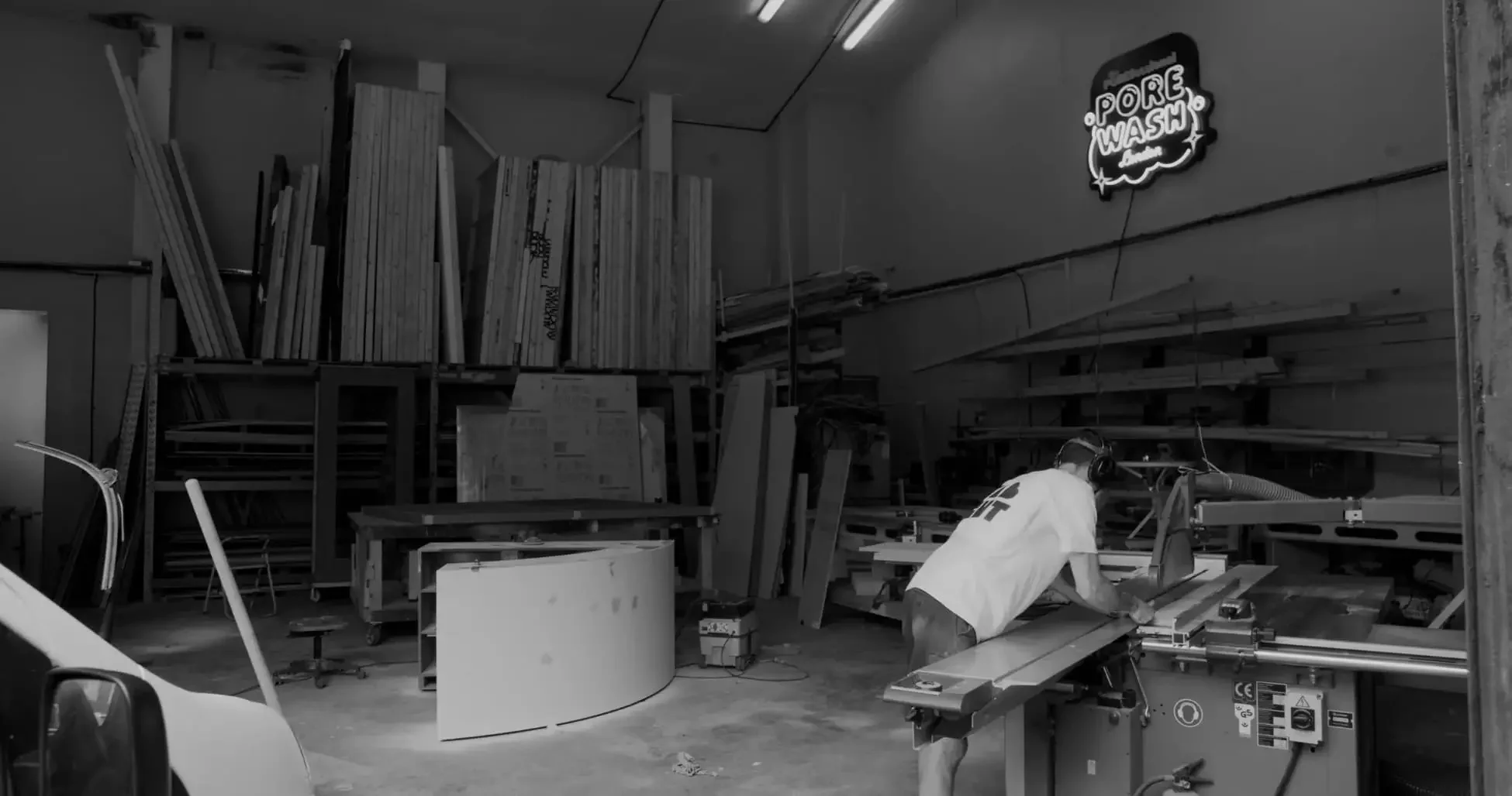 Inside a woodworking workshop with assorted wood planks stored on shelves, and a person wearing protective gear operating a table saw, with a neon sign on the wall that says 'Pore Wash'.