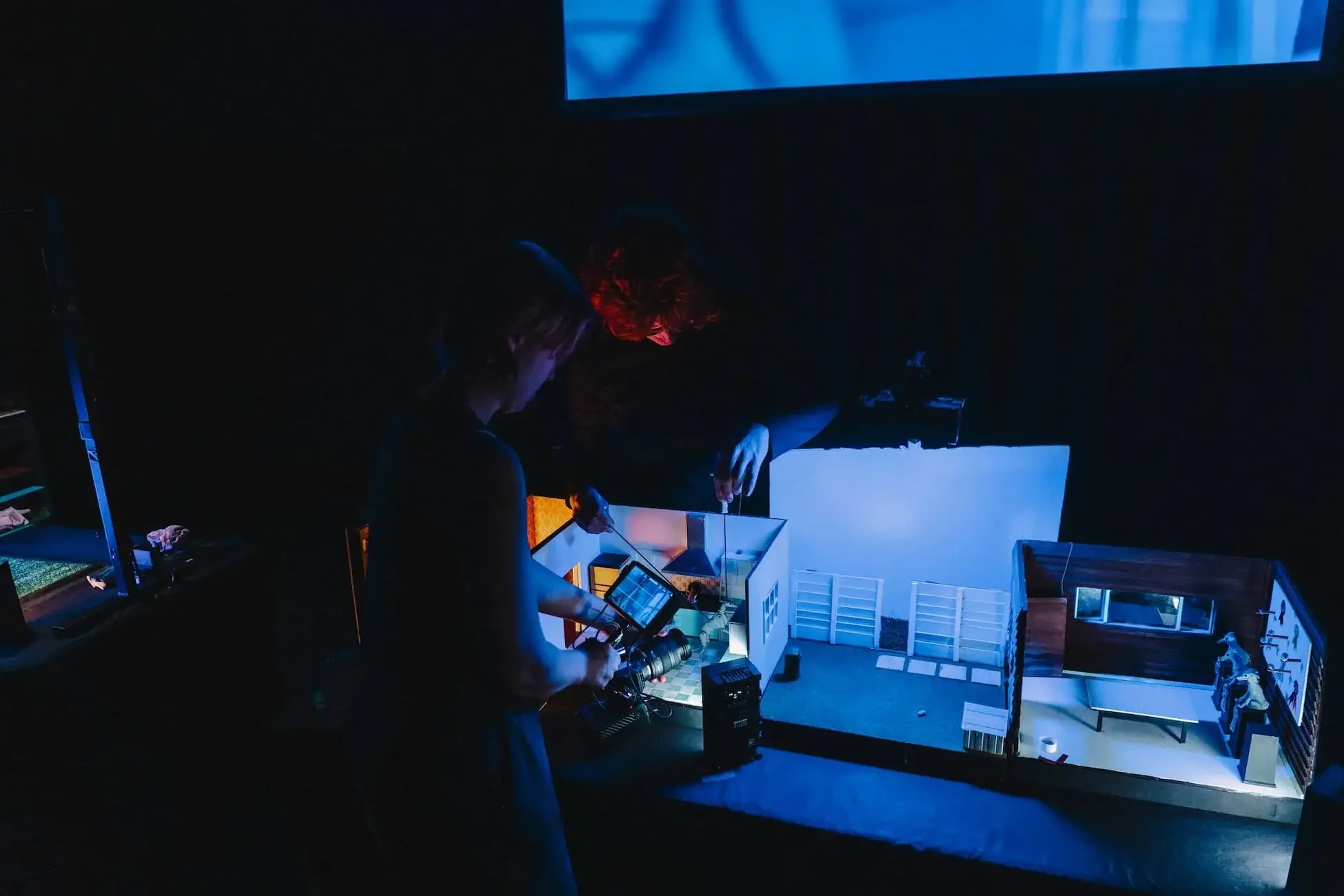 Two people working on a project in a dark room with blue lighting, surrounded by miniature house models and computer screens.