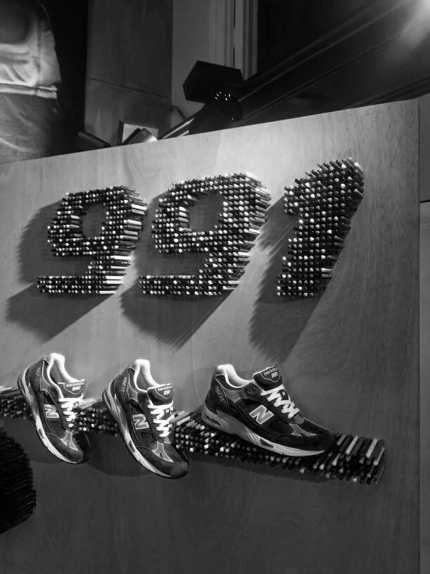 New Balance sneakers displayed on a shelf with large, decorative letters 'E&J' made of small metallic spheres on a wooden background.