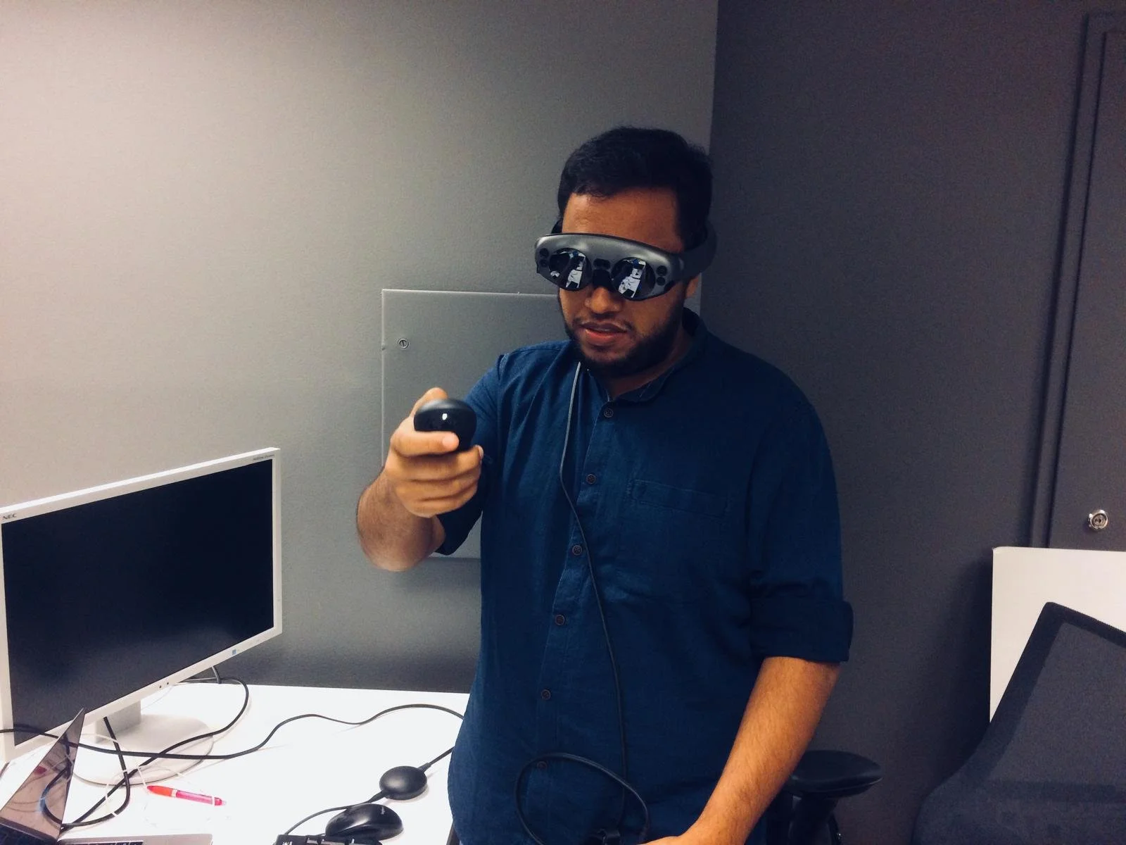A man wearing virtual reality glasses, holding a controller in his right hand, standing in an office setting with computer monitor and various cables on the desk.