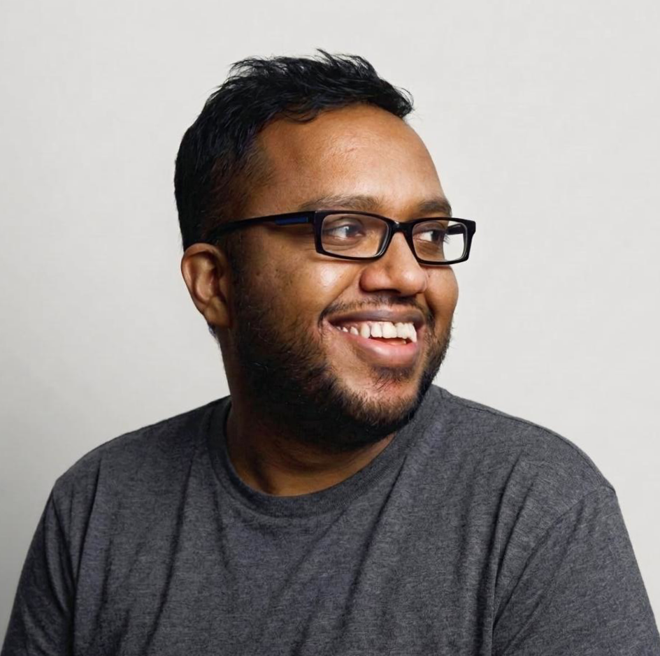 A smiling man with glasses and a beard, wearing a dark gray T-shirt, looking to the right, with a plain light gray background.