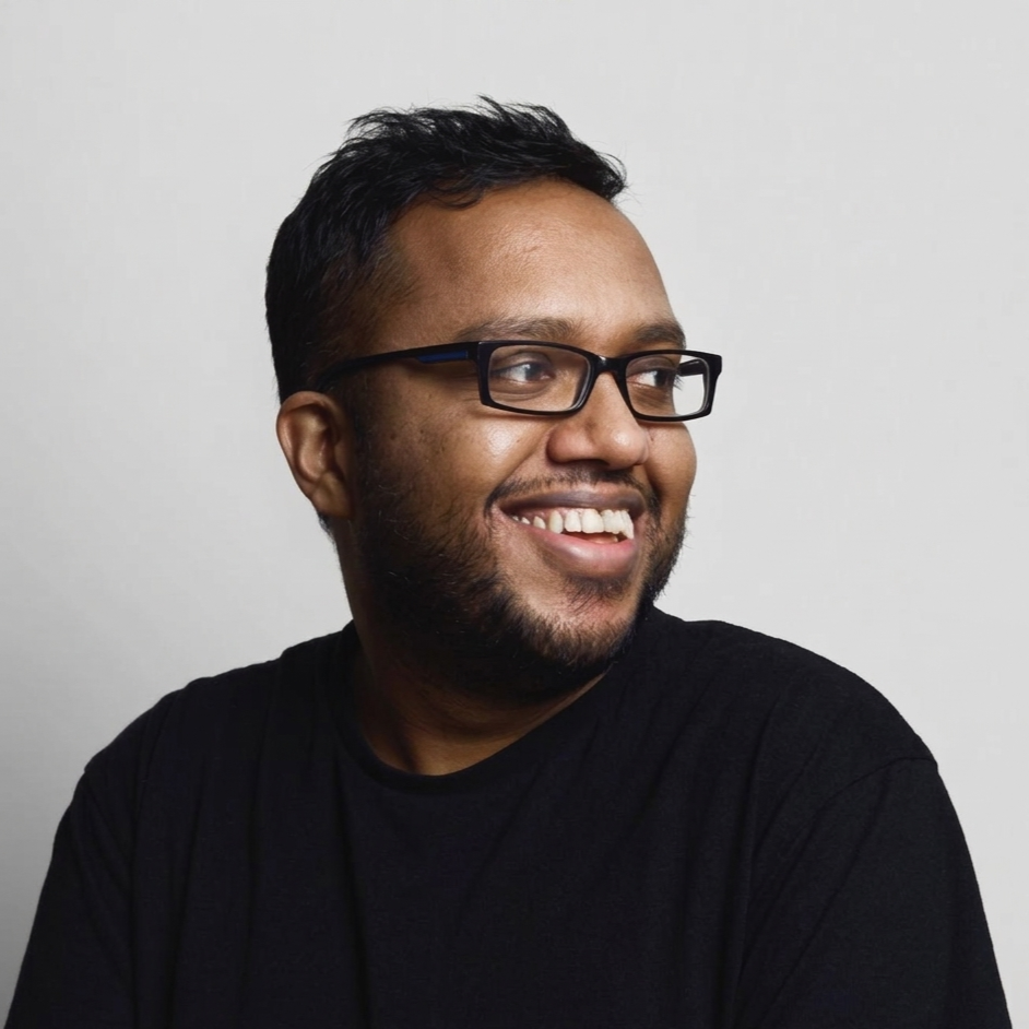 A smiling man with glasses and a beard, wearing a black shirt, looking to the side against a plain light background.