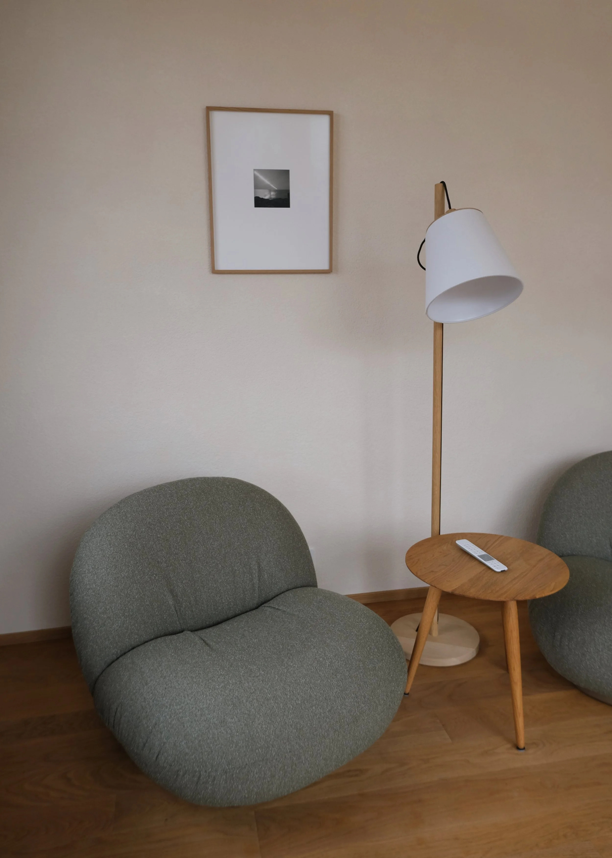 Ein sitzender, runder Stuhl mit grüner Stoffbezug, ein kleiner Holztisch mit einer Fernbedienung darauf, eine Stehlampe mit weißen Lampenschirm, eine weiße Wand mit einem eingerahmten Schwarz-Weiß-Foto.