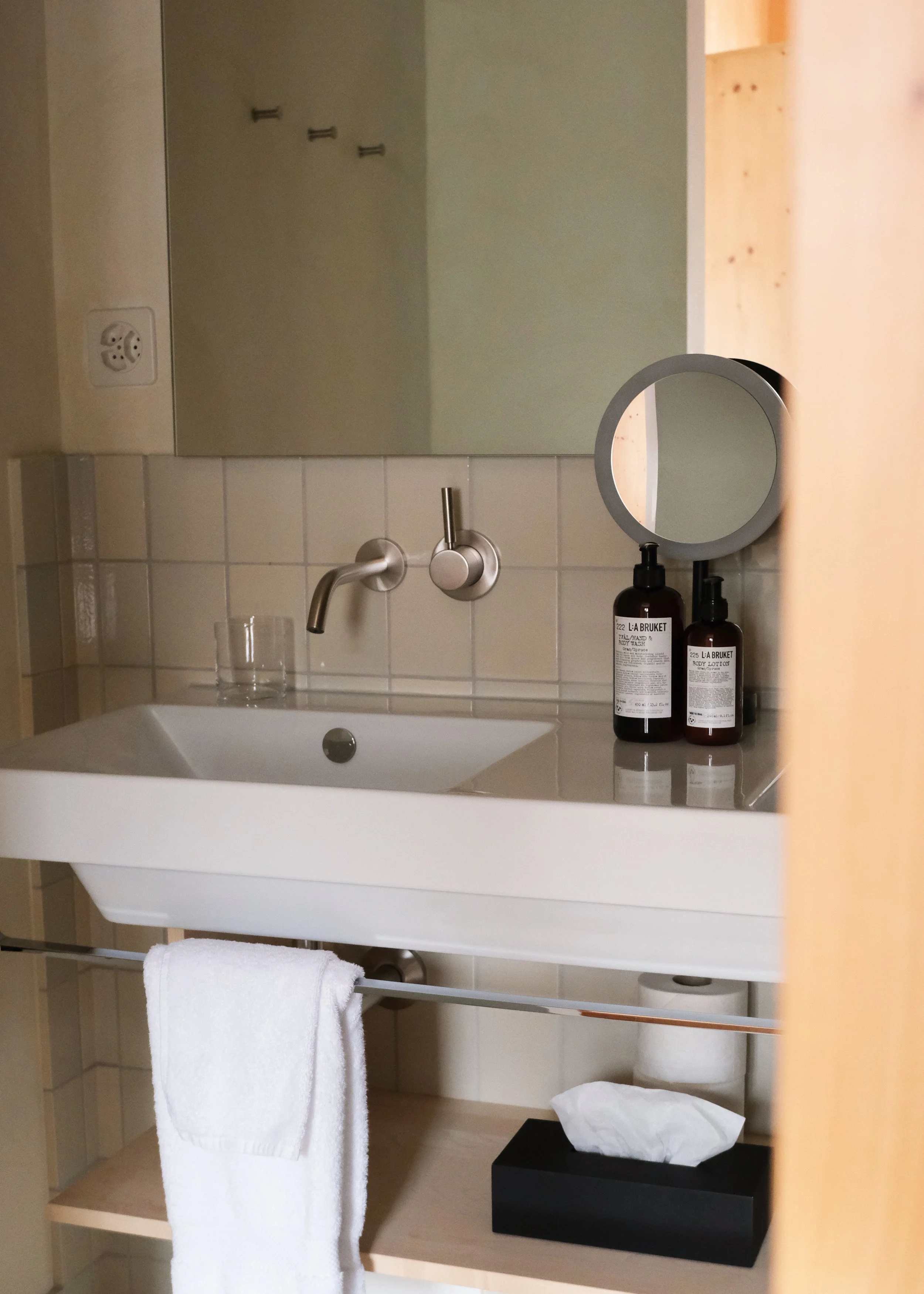 Ein Badezimmer mit einem weißen Waschbecken, einem großen Spiegel, zwei schwarzen Lotionflaschen, einem Spiegel mit Licht, einem weißen Handtuch, einem schwarzen Wäschetuch und Papierhandtüchern