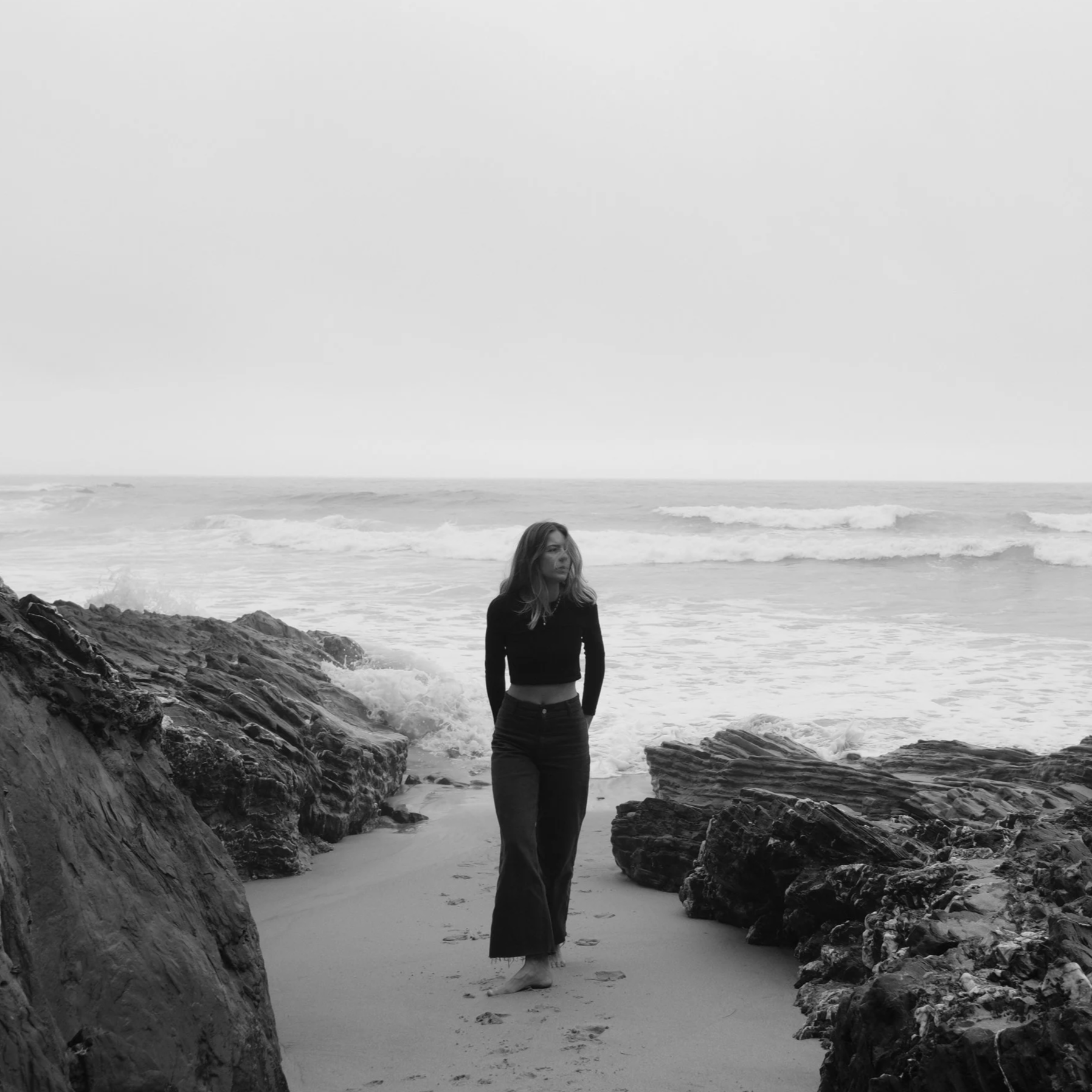 Junge Frau in schwarzen Kleid läuft barfuß am Strand entlang mit Felsen im Hintergrund, Blick aufs Meer, Schwarz-Weiß-Foto.