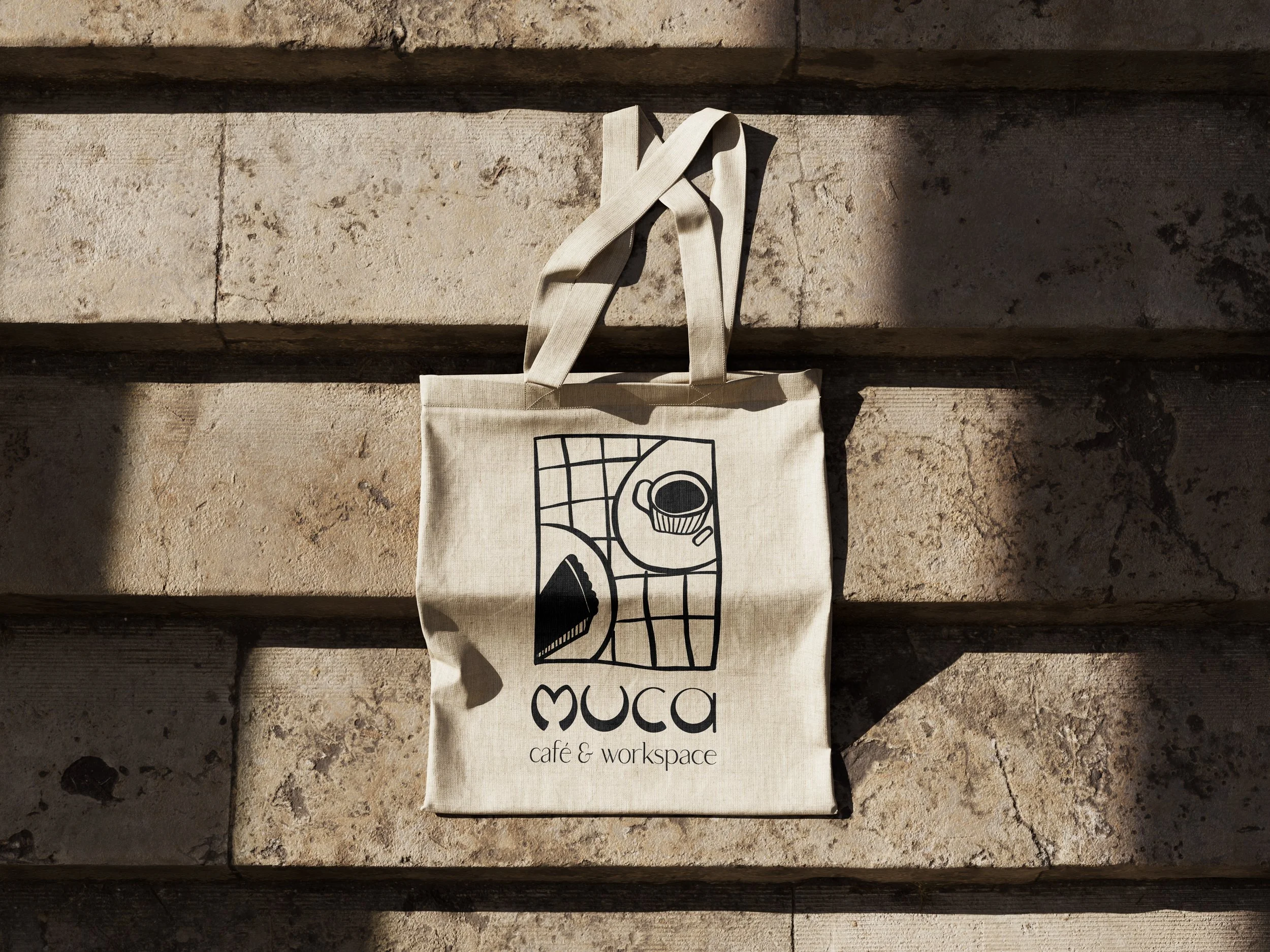 Naturfarbene Stofftasche mit schwarz-weißem Druck, auf Treppenliege liegend, mit Motto 'MUCA café & workspace'.
