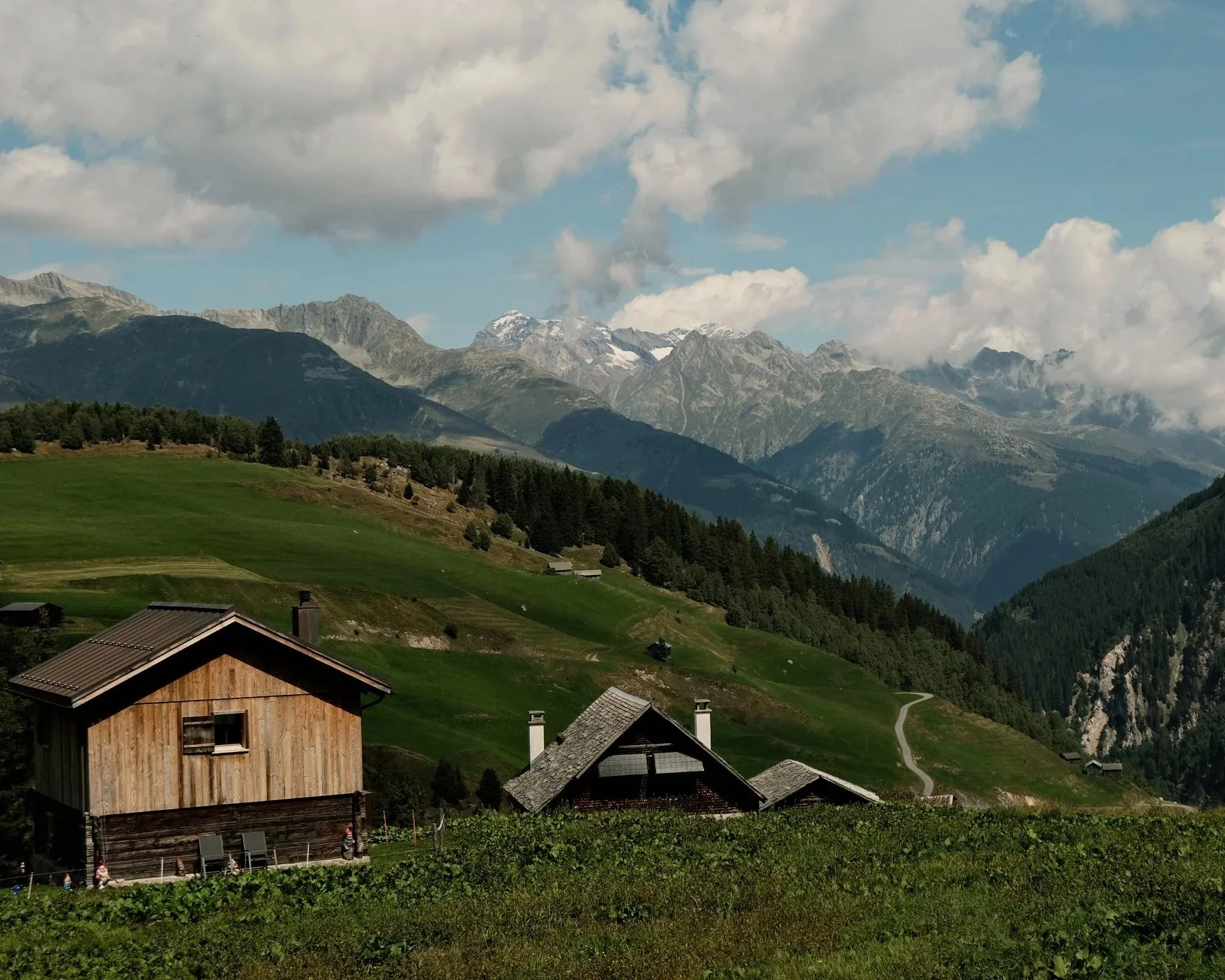 Landschaft mit grünen Hügeln, Holzhaus und Bergkette im Hintergrund.