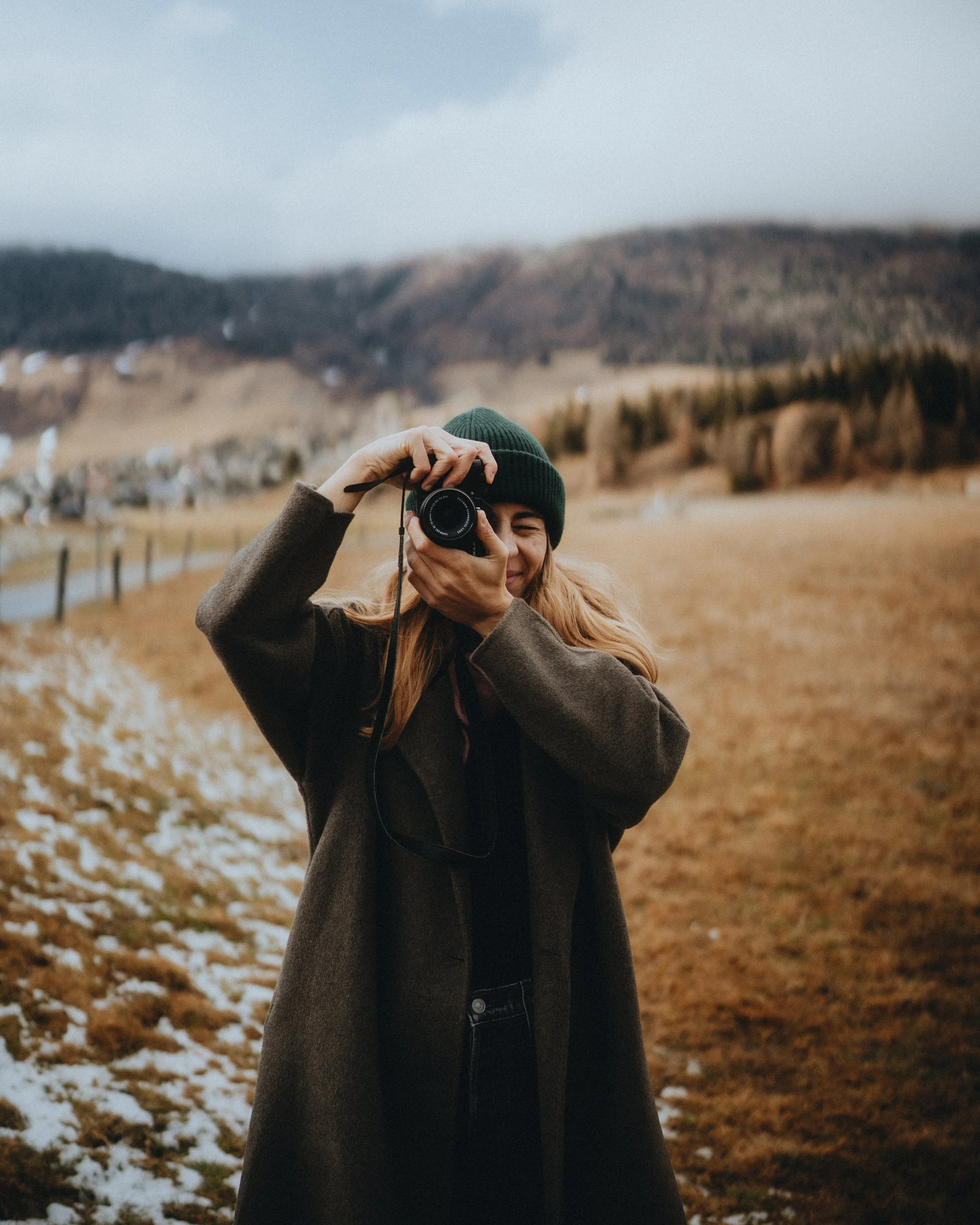 Eine lachende Frau mit braunem Mantel und grüner Mütze fotografiert mit einer Kamera in einer ländlichen, bergigen Landschaft bei bedecktem Himmel.