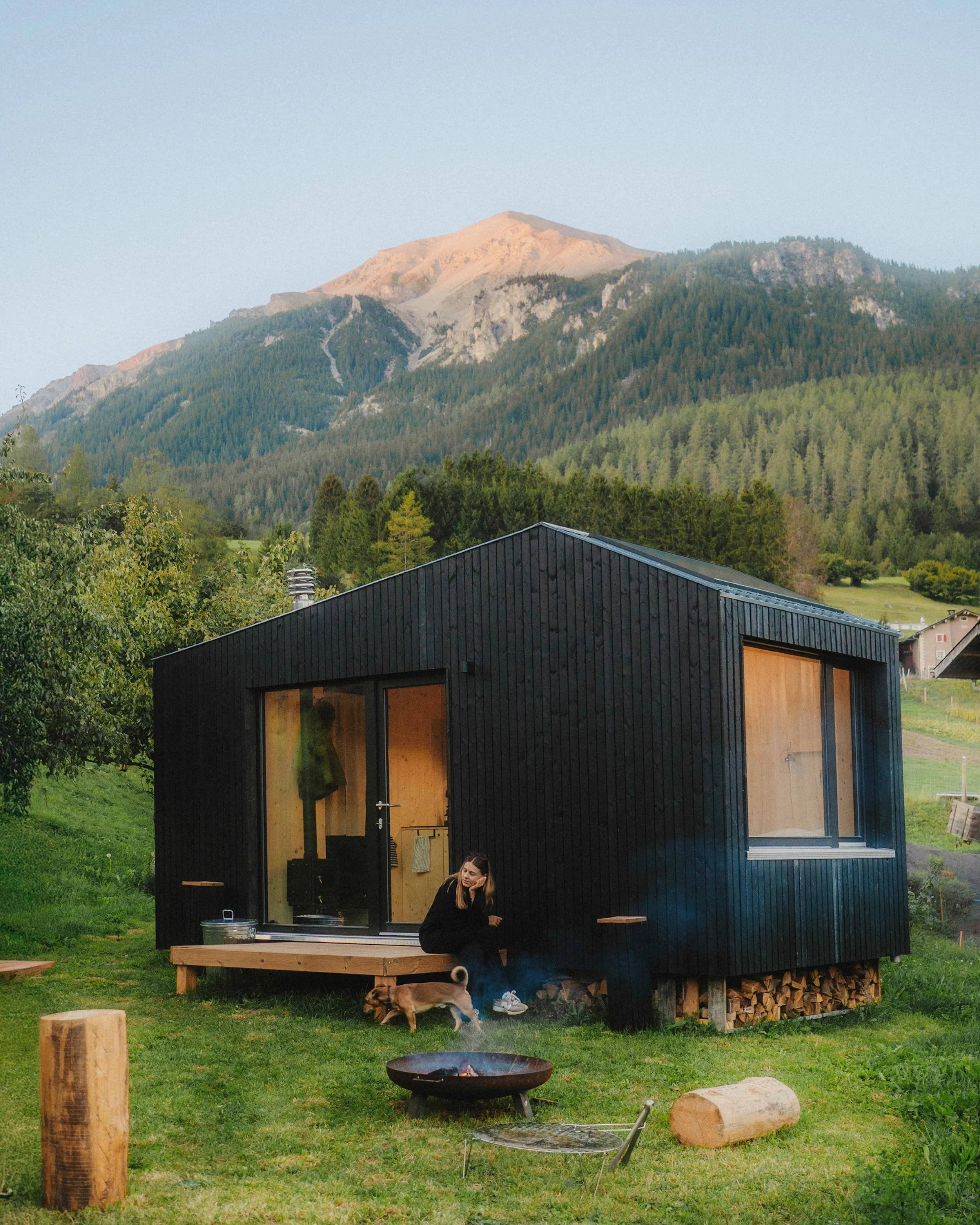 Eine schwarze moderne Hütte mit großen Fenstern in einer grünen, bergigen Landschaft, ein Mensch sitzt vor der Hütte mit einem Hund, im Vordergrund ein Feuerkorb und Holzstämme.