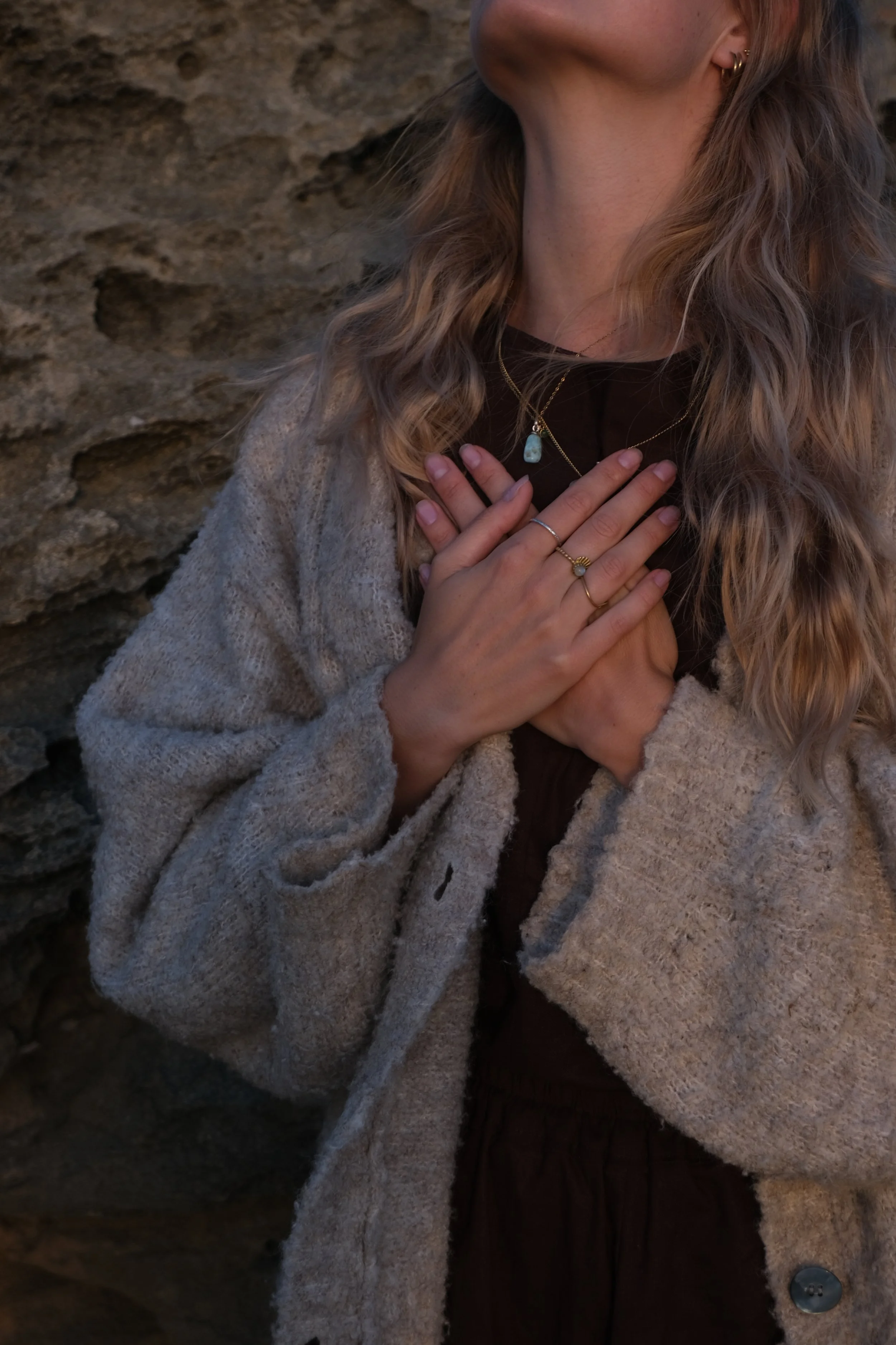 Nahaufnahme eines Frauenhändes, die die Brust berührt, Steine im Hintergrund, getragenes Schmuck, lockiges Haar, beige Jacke.