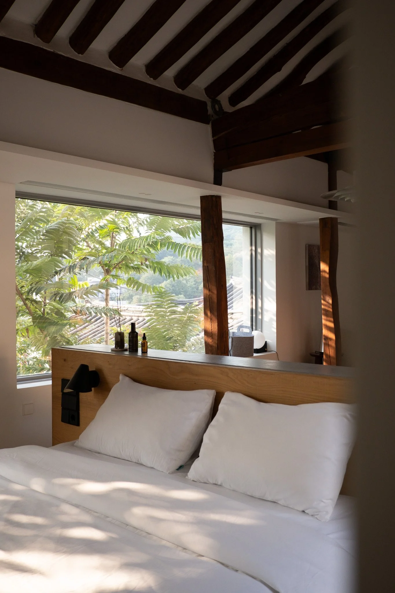 Bett mit weißen Kissen in einem modernen Schlafzimmer mit großen Fenstern und Blick auf grüne Bäume, Holzdekor und natürlichen Lichtverhältnissen.