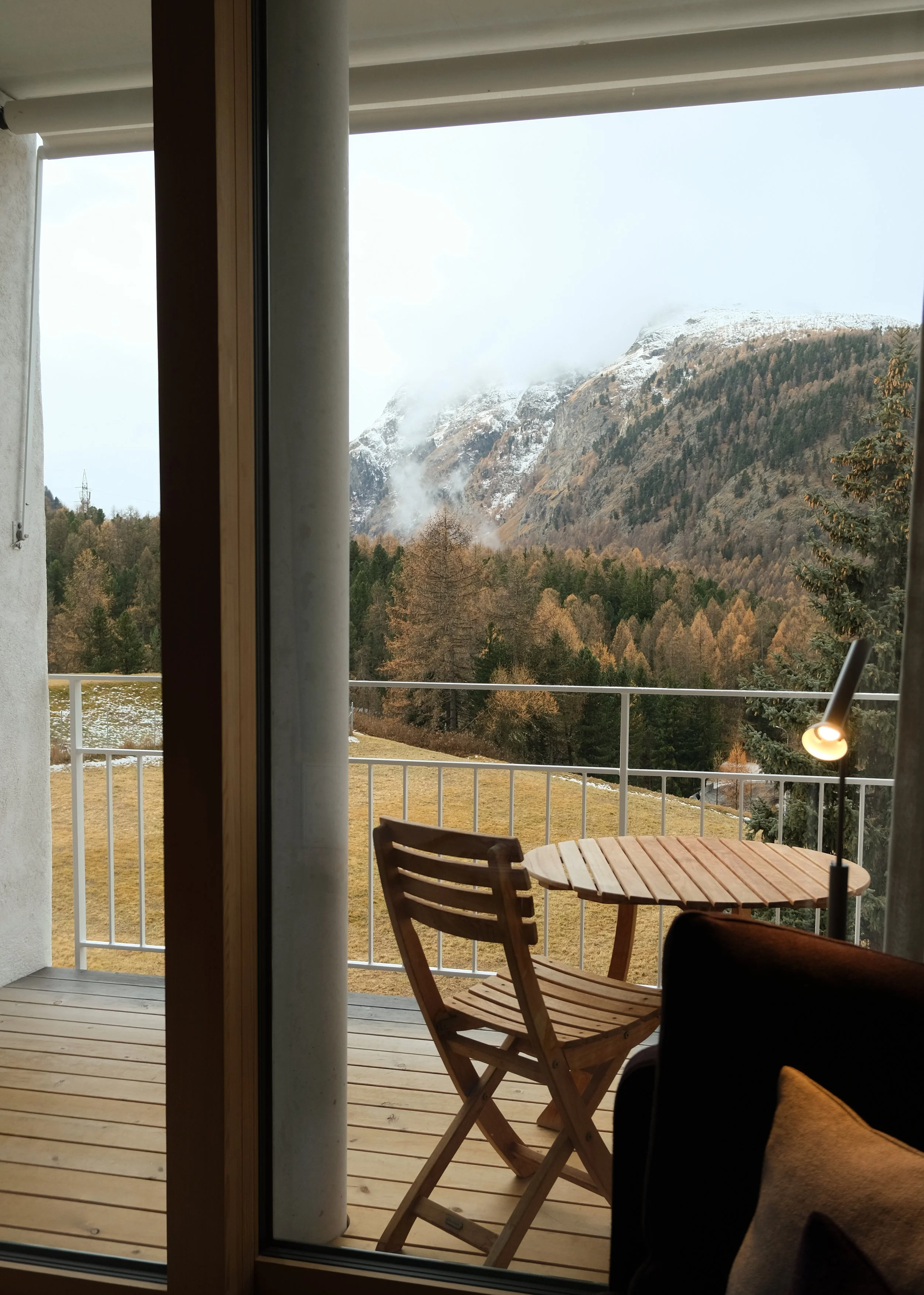 Balkon mit einem Holzstuhl und einem Tisch, Blick auf eine bergige Landschaft mit Bäumen und verschneiten Gipfeln im Hintergrund.