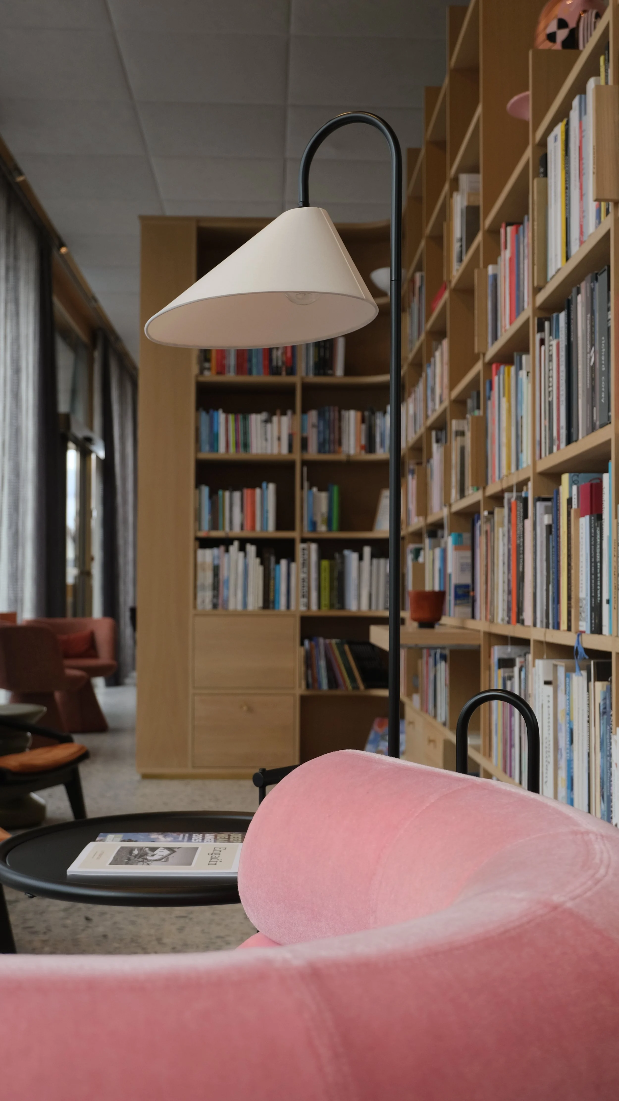 Ein Sicht auf eine gemütliche Leseecke mit einem pinken Sessel, einem schwarzen Beistelltisch mit einem Buch, einen Stehlampe mit cremefarbenem Lampenschirm und eine große Bücherwand im Hintergrund.