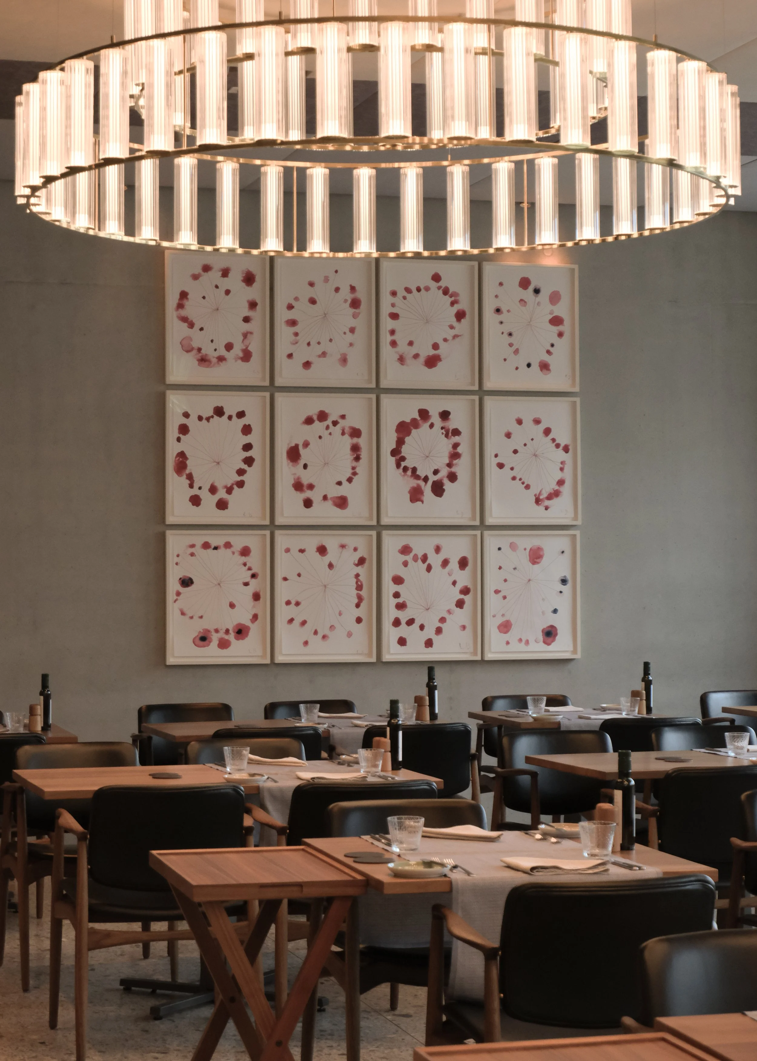 Ein stilvoller Restaurantraum mit einem großen, modernen Kronleuchter, der aus vielen vertikalen Leuchten besteht, und einer Wand mit neun quadratischen Kunstwerken, die botanische Illustrationen von Blüten in Rot- und Pinktönen zeigen.