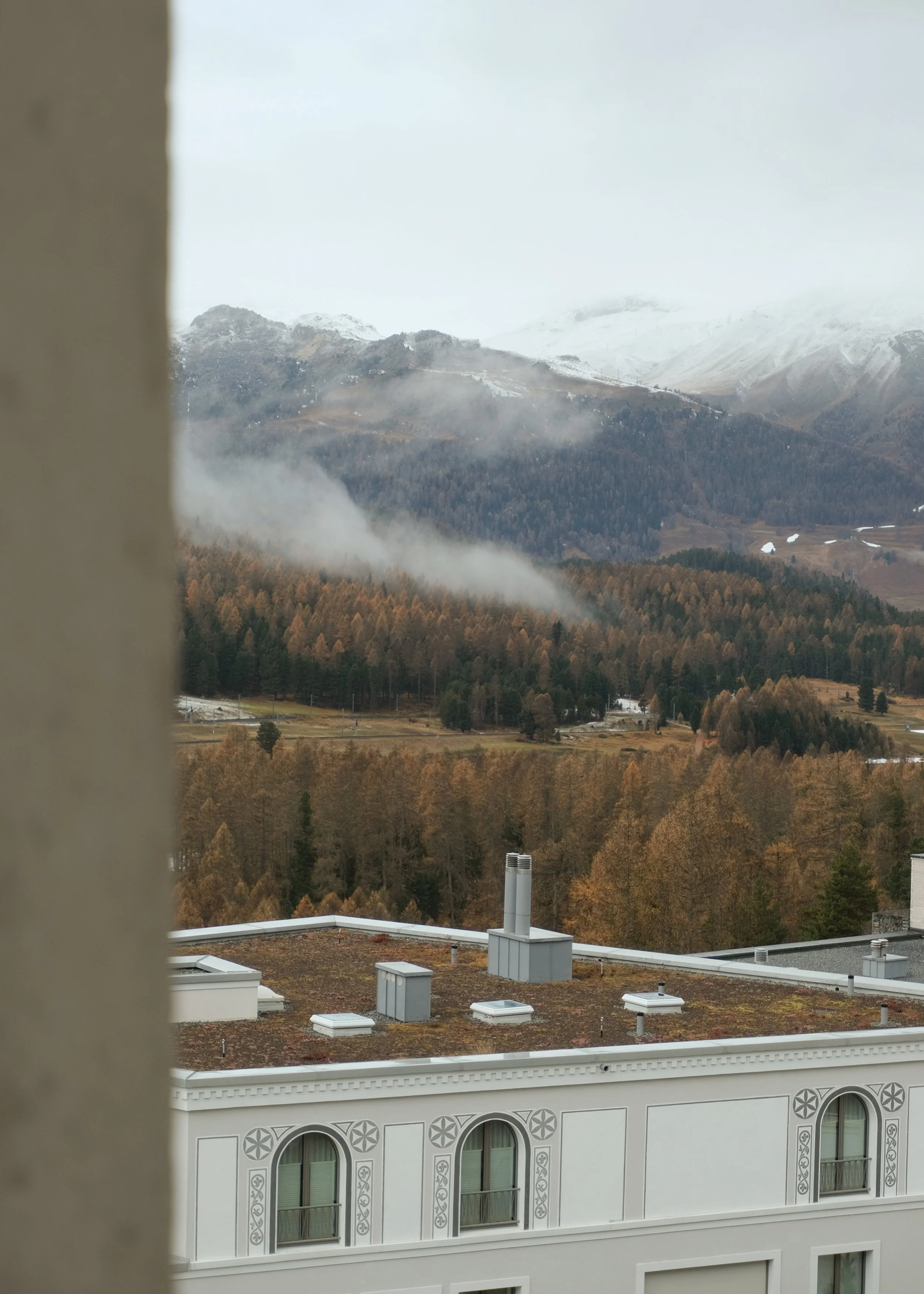 Blick auf ein Gebäude mit weißen Wänden und dekorativen Fenstern im Vordergrund, im Hintergrund eine bergige Landschaft mit Wäldern, Wolken und Nebel, sowie schneebedeckte Berge.