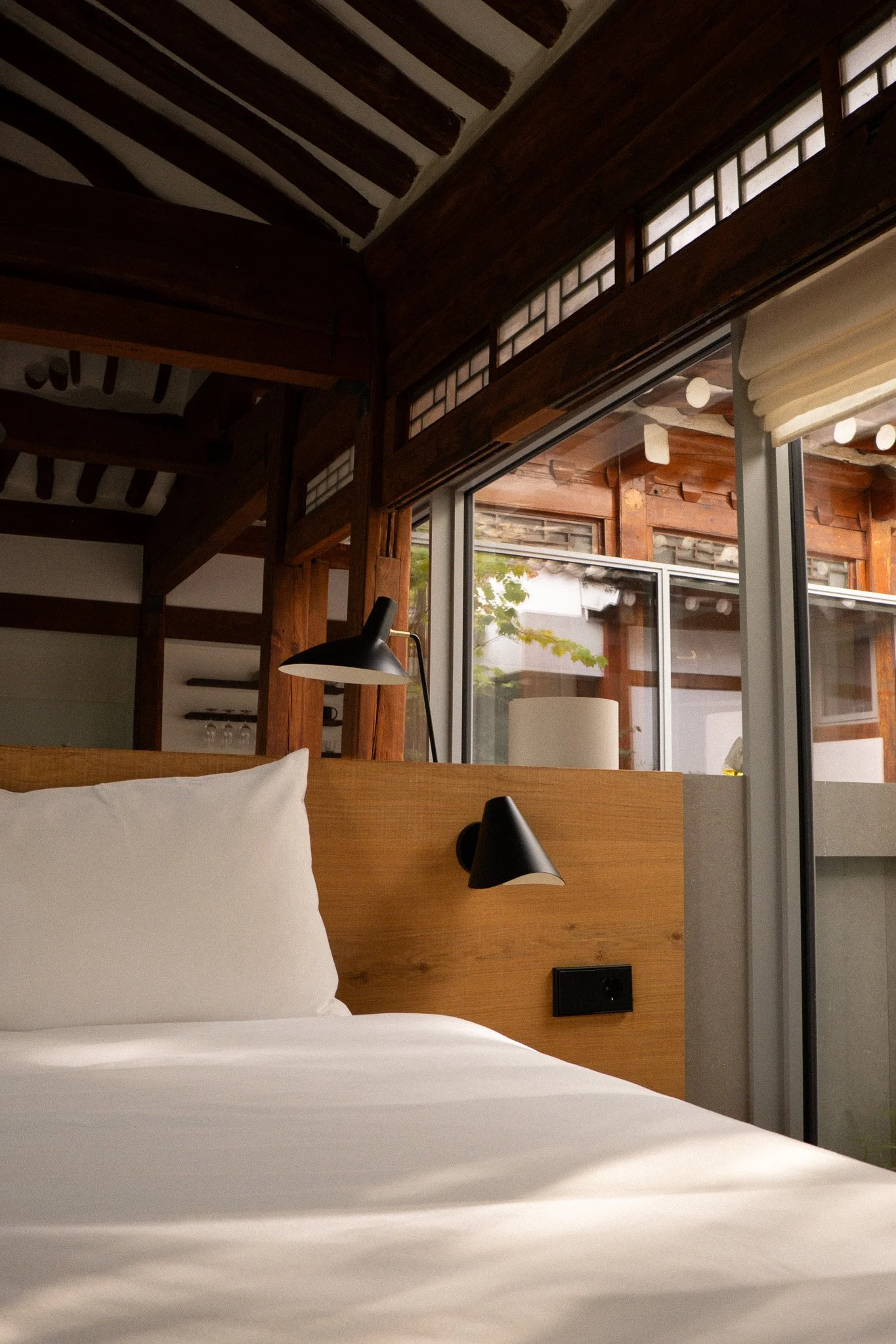 Ein Schlafzimmer mit einem karierten Bett, einer Holzwand, einem Nachttisch, einer schwarzen Lampe und großen Fenstern mit Sicht auf einen Außenbereich mit Holzverkleidung und Pflanzen.
